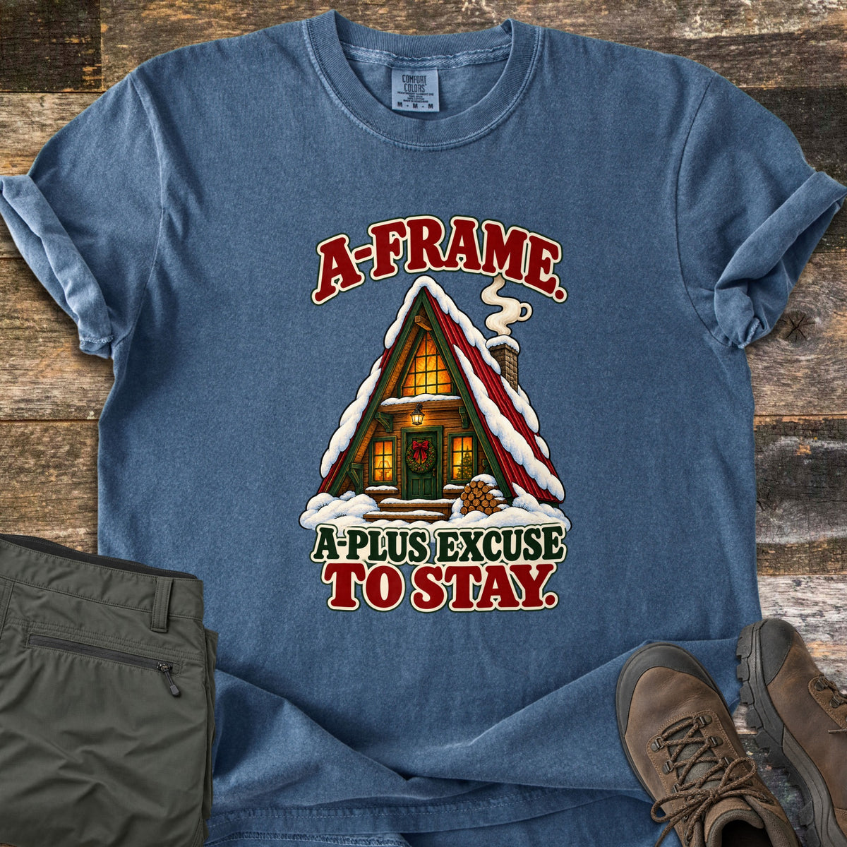 A-Frame A-Plus Excuse to Stay Comfort Colors T-shirt in Denim, laid flat. A snow-covered A-frame cabin with a red roof, glowing windows, a wreath on the door and a smoking chimney sits between the words A-Frame. and A-Plus Excuse to Stay. Olive pants and brown boots lie beside it on rustic wood.