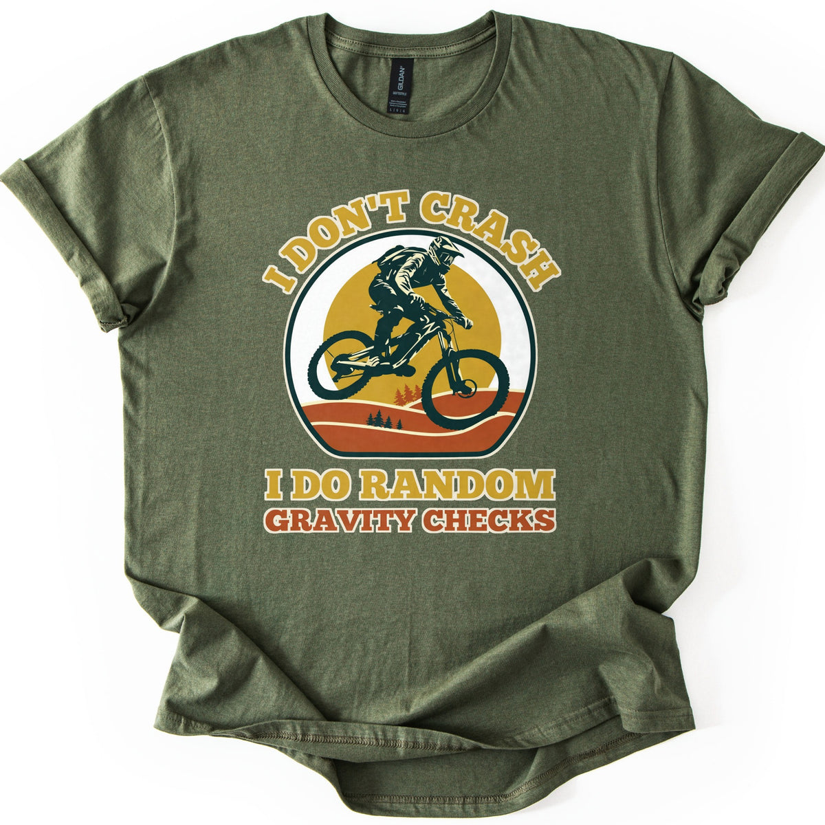 Random Gravity Checks MTB Comfort Colors T-shirt in Military Green, laid flat. A mountain biker in a full-face helmet jumps against a mustard sun over orange hills with pine trees, framed by the words I Don't Crash above and I Do Random Gravity Checks below. Sleeves rolled, on white.