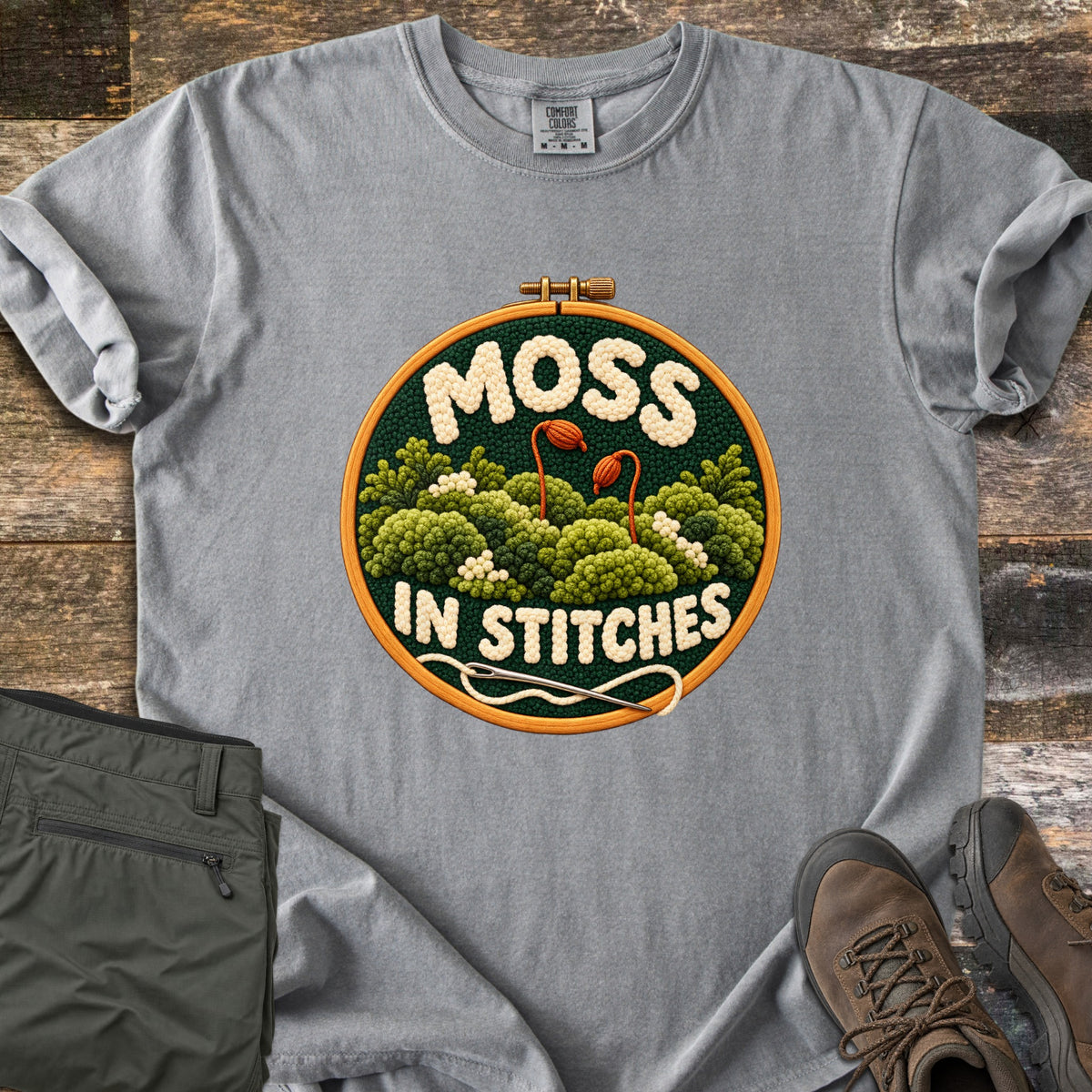 Moss in Stitches Comfort Colors T-shirt in Grey, laid flat. An embroidery hoop holds stitched green moss with two orange spore capsules, the word Moss above and In Stitches below, plus a needle and thread. Folded olive pants and brown hiking boots lie beside it on weathered wood.
