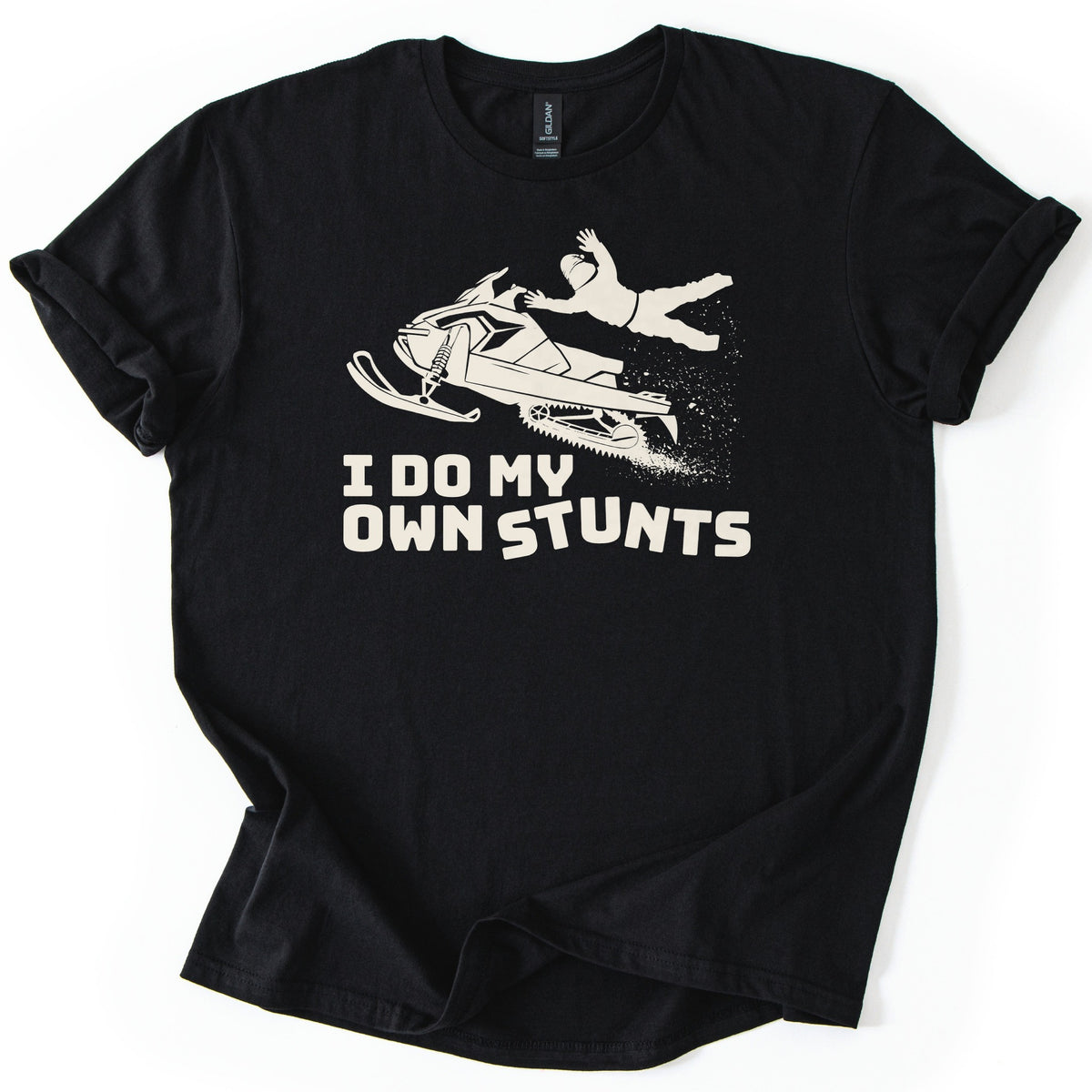 Snow Mobile Stunts Comfort Colors T-shirt in Black, laid flat. A cream silhouette of a snowmobile flying through the air, its rider thrown off behind it in a spray of snow, above the words I Do My Own Stunts. The shirt lies on a white background with rolled sleeves.