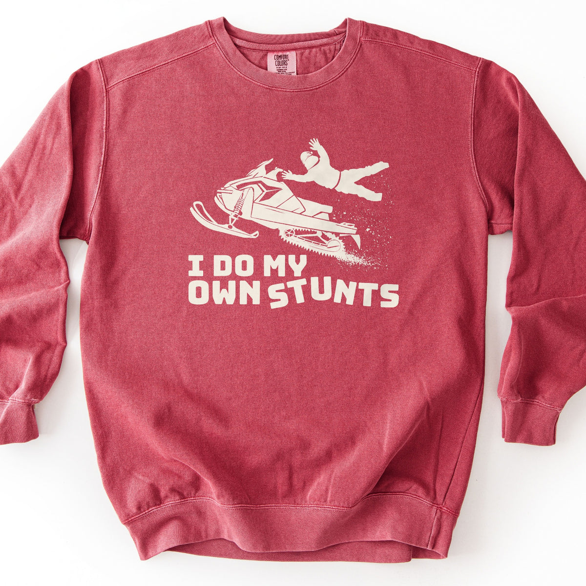 I Do My Own Stunts Comfort Colors crewneck sweatshirt in Crimson, laid flat. A cream print shows a rider in a helmet tossed off a snowmobile in midair as snow sprays out, above the words I DO MY OWN STUNTS. The sweatshirt lies on a white surface.