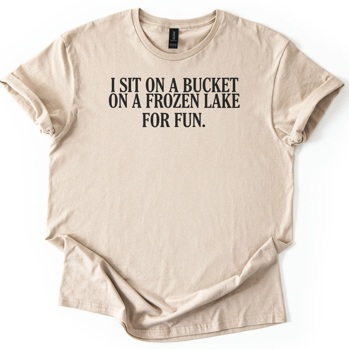 Bucket On A Frozen Lake Comfort Colors T-shirt in Sand, laid flat. Black serif capital letters across the chest read I SIT ON A BUCKET ON A FROZEN LAKE FOR FUN. The shirt lies on a white surface with its sleeves rolled and the hem loosely folded.