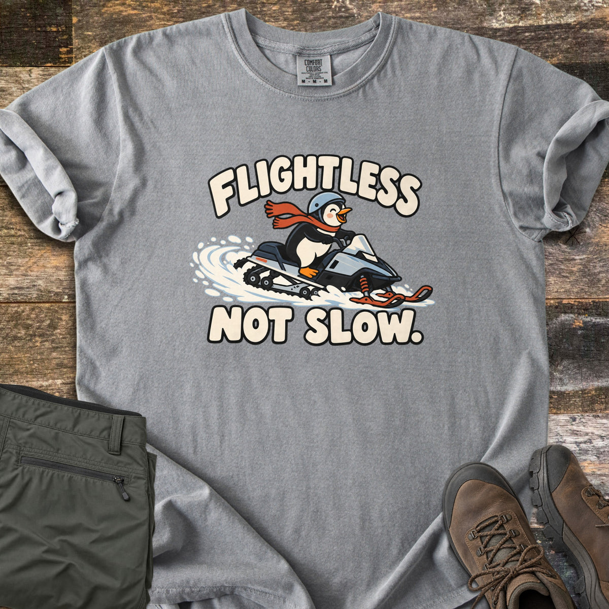 Flightless Not Slow Comfort Colors T-shirt in Grey, laid flat. A cartoon penguin in a helmet and red scarf rides a snowmobile through snow between the words Flightless Not Slow. Folded olive pants and brown hiking boots sit beside it on weathered wood.