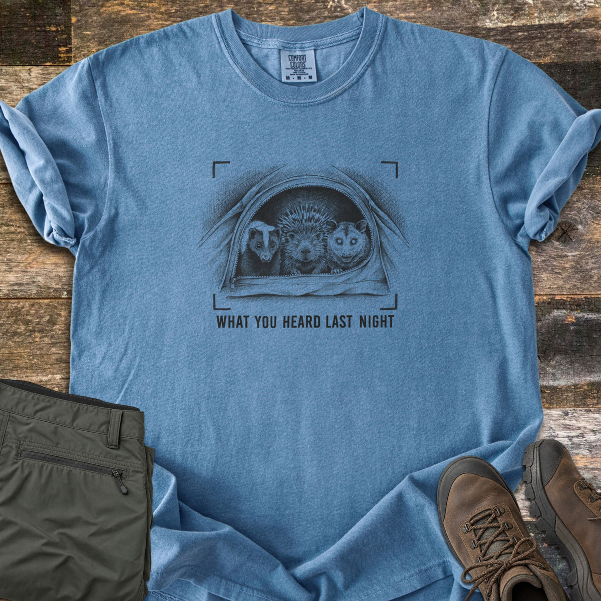 What You Heard Last Night Comfort Colors T-shirt in Blue Jean, laid flat. A black sketch of a skunk, porcupine and opossum peeking out of an unzipped tent, framed by camera brackets, above the words What You Heard Last Night. Olive hiking pants and brown boots lie on weathered wood.