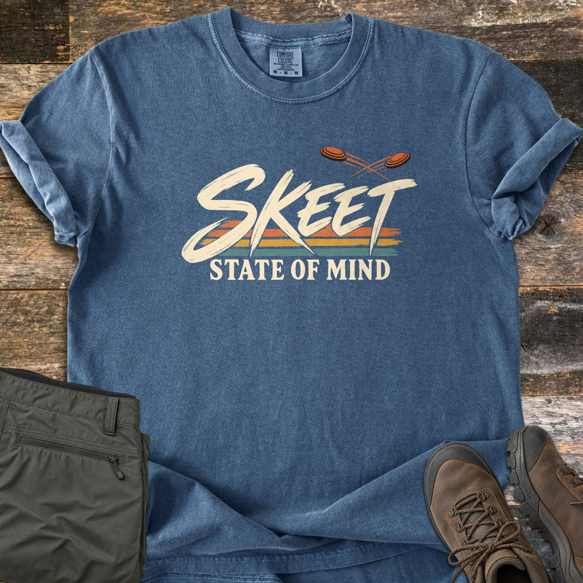 Skeet State of Mind Comfort Colors T-shirt in Denim, laid flat. Brush-script SKEET over orange, yellow and teal stripes with two flying clay targets, and STATE OF MIND below. Olive cargo pants and brown hiking boots sit beside it on weathered wood planks.
