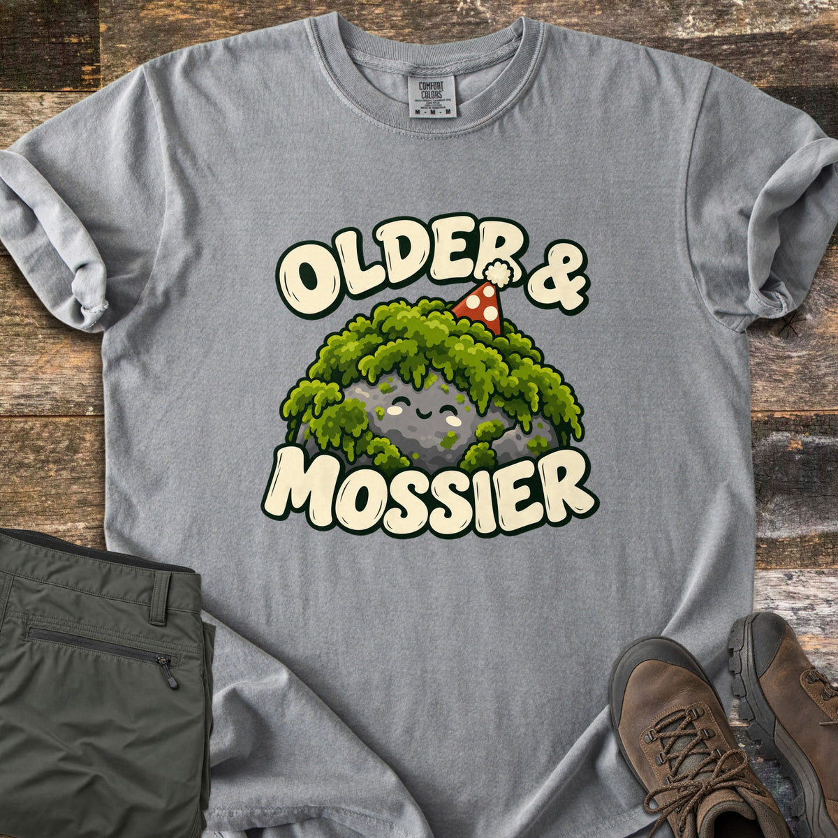 Older & Mossier Comfort Colors T-shirt in Grey, laid flat. The words Older & Mossier in cream letters surround a smiling gray rock covered in green moss, wearing a red polka-dot party hat. Folded olive hiking pants and brown hiking boots sit on weathered wood.