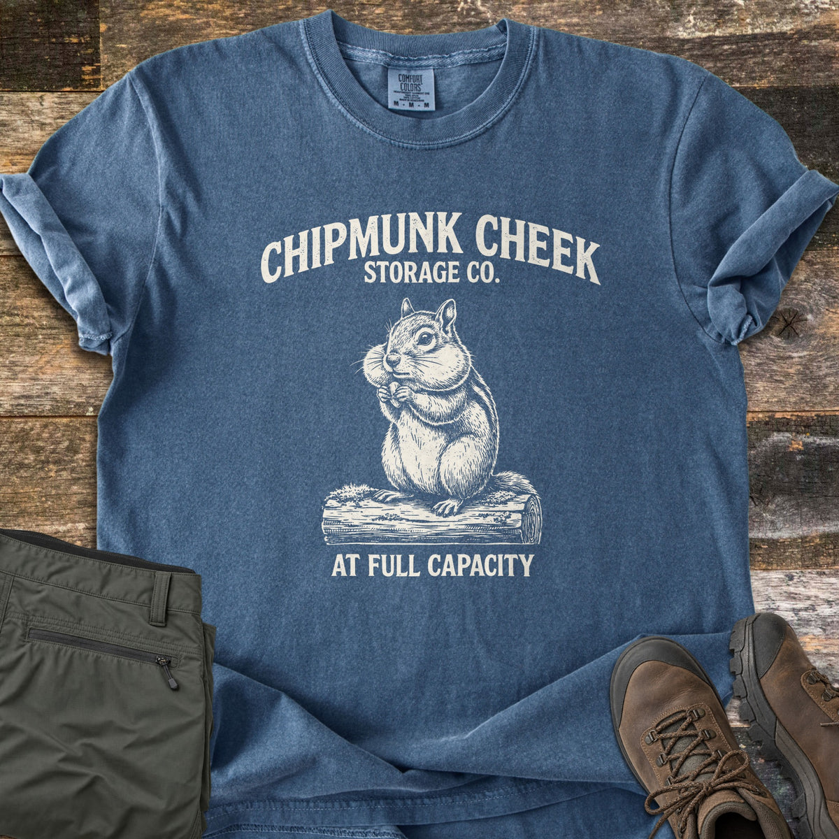 Chipmunk Cheek Storage Co Comfort Colors T-shirt in Denim, laid flat. A vintage-style sketch of a chipmunk with full cheeks sitting on a log, under the words Chipmunk Cheek Storage Co. and above At Full Capacity. Shown on weathered wood with olive hiking pants and brown hiking boots.
