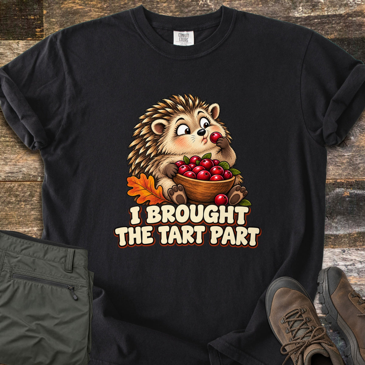 I Brought the Tart Part Comfort Colors T-shirt in Black, laid flat. A cartoon hedgehog nibbles a cranberry while holding a wooden bowl of cranberries beside an orange oak leaf, above the words I Brought the Tart Part. Olive pants and brown hiking boots sit on a wood background.