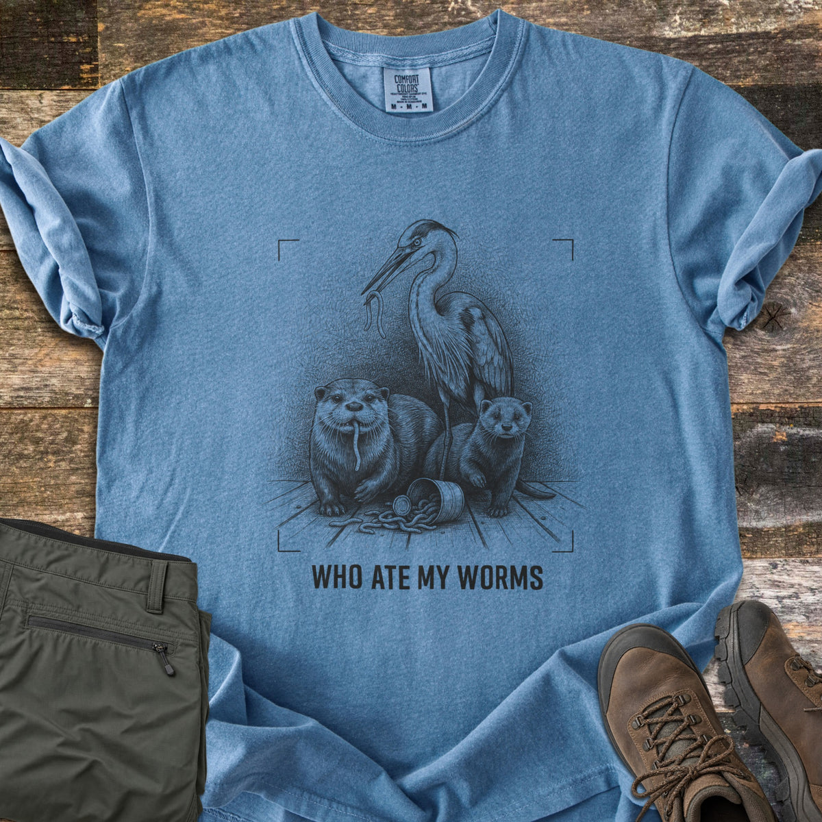 Who Ate My Worms Comfort Colors T-shirt in Blue Jean, laid flat. A black sketch of a heron and an otter each eating a worm beside a mink and a spilled bait can, inside viewfinder corners, above the words Who Ate My Worms. Olive pants and brown hiking boots lie on weathered wood.