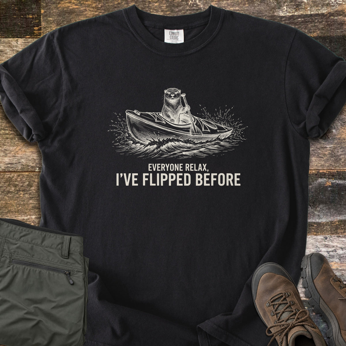 Everyone Relax I've Flipped Before Comfort Colors T-shirt in Black, laid flat. A cream sketch of an otter paddling a kayak through splashing waves above the words Everyone Relax, I've Flipped Before. Sleeves are rolled, with olive hiking pants and brown boots on rustic wood planks.