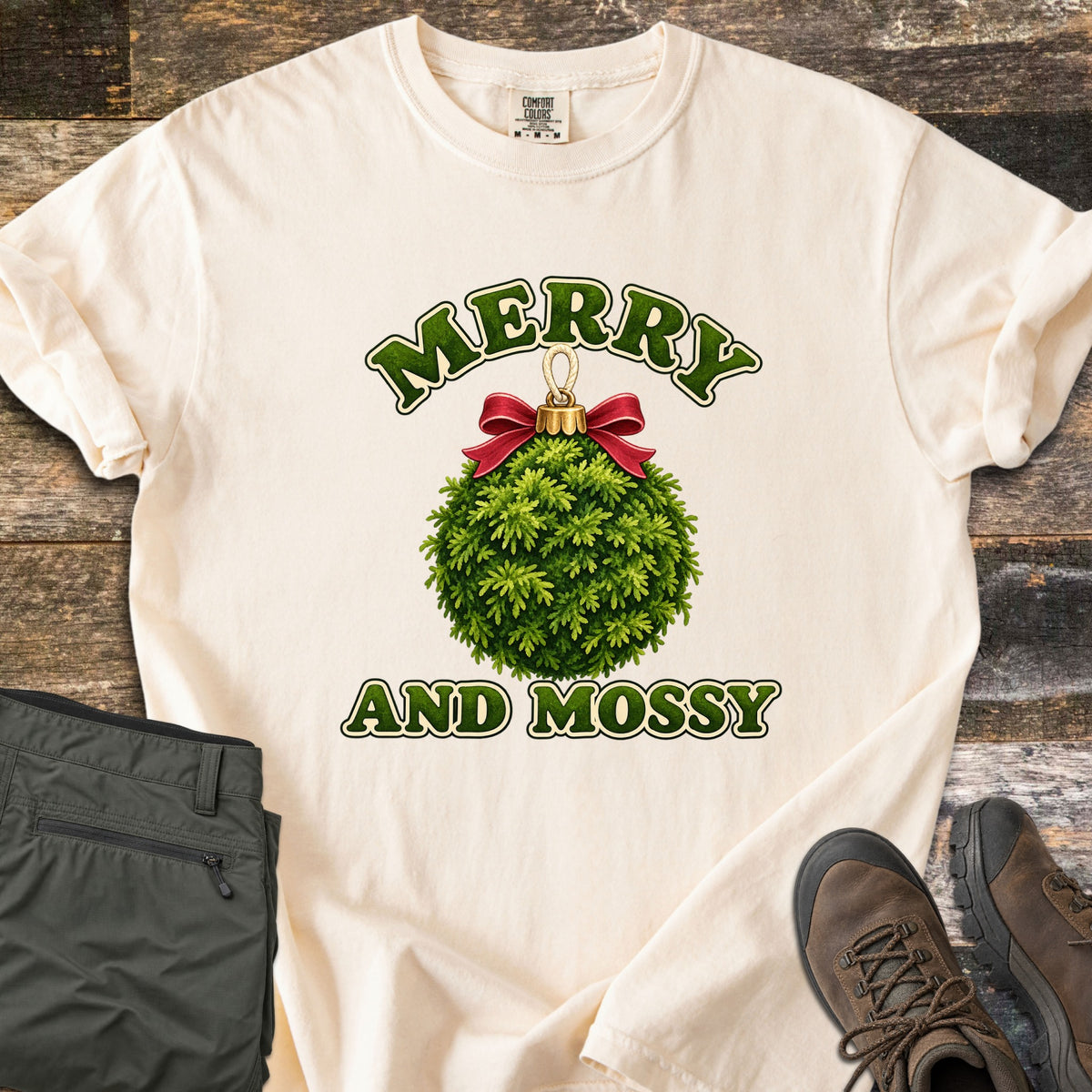 Merry and Mossy Comfort Colors T-shirt in Ivory, laid flat. A round Christmas ornament made of green moss, topped with a gold cap and red bow, sits between the words Merry and Mossy in green lettering. Olive hiking pants and brown hiking boots lie beside it on weathered wood.