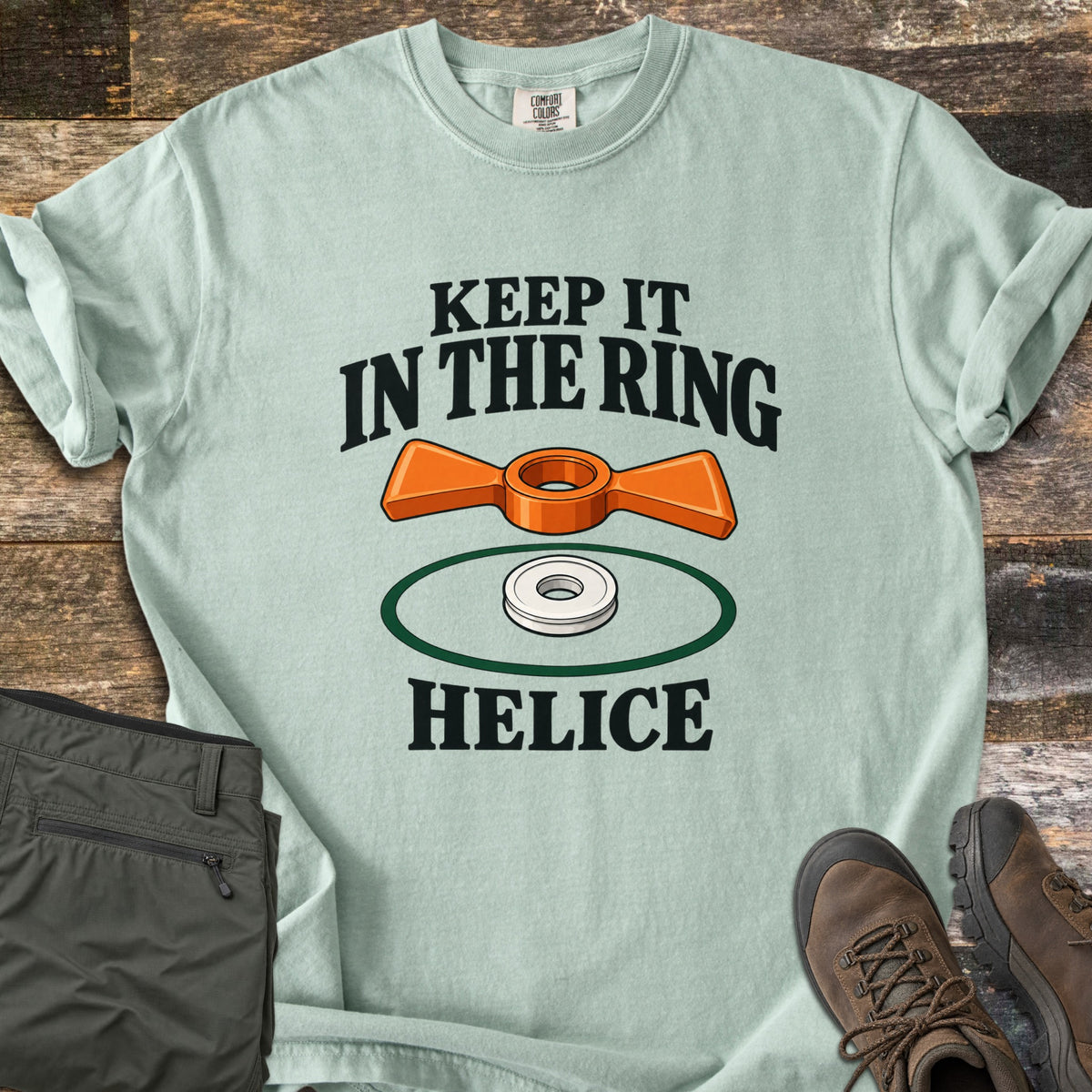 Keep It in the Ring Comfort Colors T-shirt in Bay, laid flat. An orange winged helice target floats above a white ring inside a green circle, between the words Keep It in the Ring and Helice. Olive pants and brown hiking boots sit beside it on weathered wood.