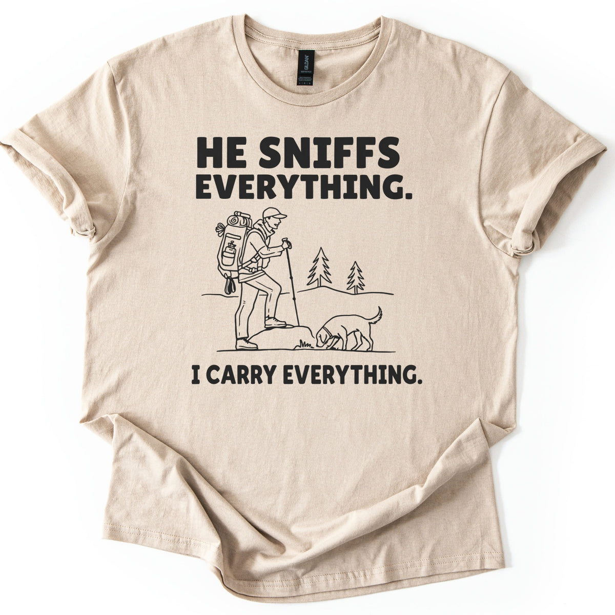 Trail Dog Pack Mule Comfort Colors T-shirt in Sand, laid flat. Black line art of a hiker with a loaded backpack and trekking pole stepping onto a rock as a dog sniffs the ground, with pine trees behind, between the words He sniffs everything. and I carry everything. Sleeves rolled, on white.