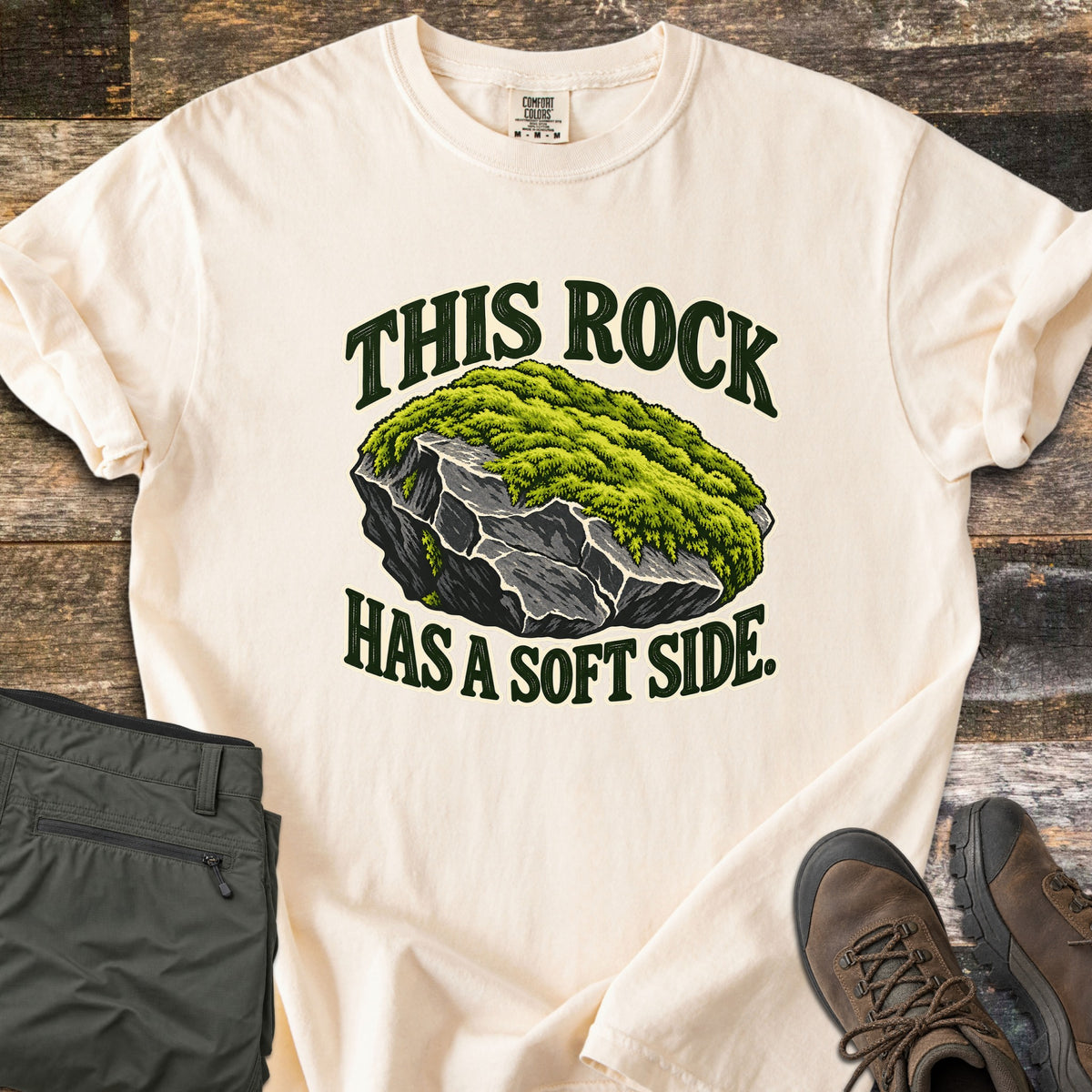 This Rock Has a Soft Side Comfort Colors T-shirt in Ivory, laid flat. An illustrated gray boulder topped with thick green moss sits between the words This Rock above and Has a Soft Side. below. Olive hiking pants and brown hiking boots lie beside it on weathered wood planks.