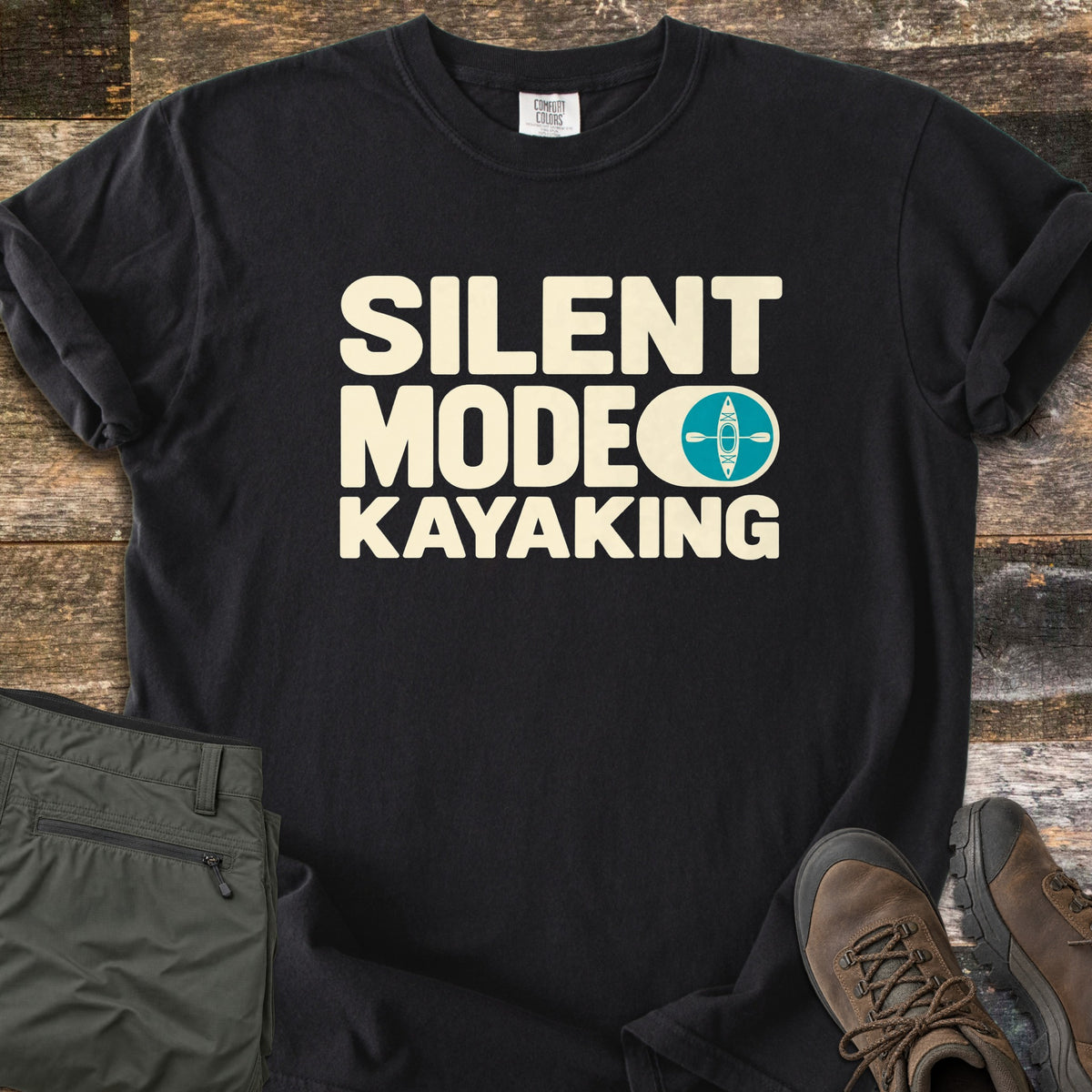 Silent Mode Comfort Colors T-shirt in Black, laid flat. Cream block letters read SILENT MODE KAYAKING, with a toggle switch icon showing a teal circle with a kayak and paddle. Sleeves are rolled on a wood background beside folded olive pants and brown hiking boots.