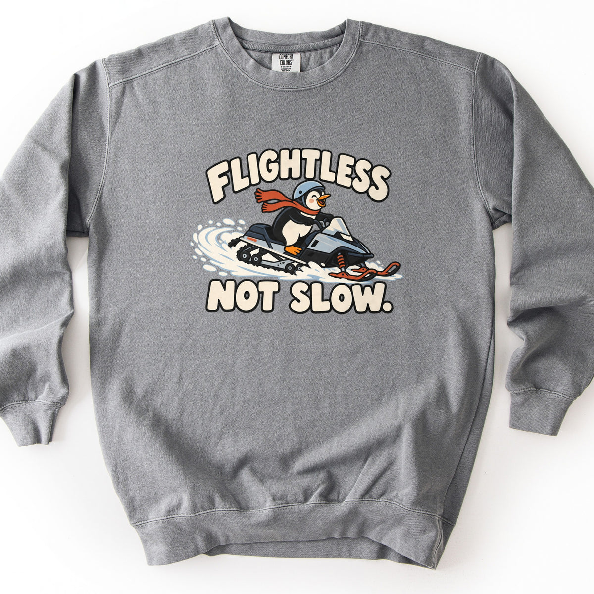 Flightless Not Slow Comfort Colors crewneck sweatshirt in Grey, laid flat. A cartoon penguin in a blue-gray helmet and red scarf rides a gray snowmobile through spraying snow, between the cream words Flightless and Not Slow. It lies on a white background.