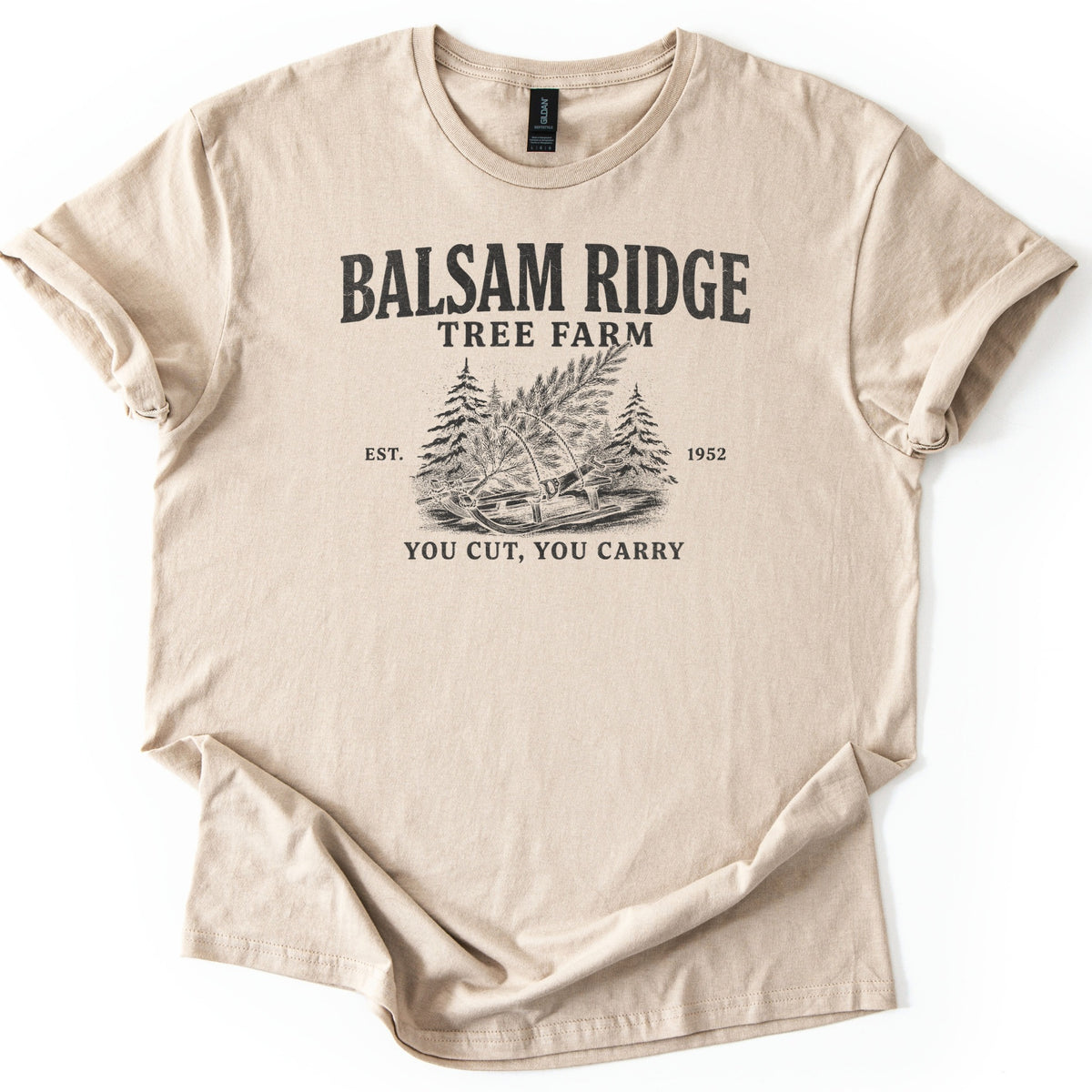 Balsam Ridge Tree Farm Comfort Colors T-shirt in Sand, laid flat. Black print reads Balsam Ridge Tree Farm above an engraved-style fir tree tied to a wooden sled between pines, flanked by Est. 1952, with You Cut, You Carry below. Sleeves cuffed on a white surface.