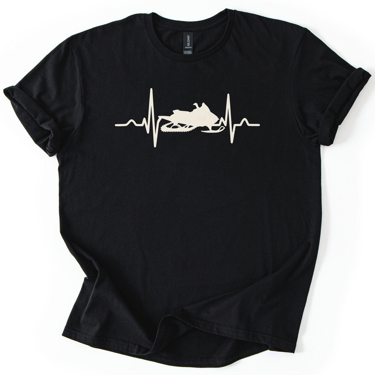 Heartbeat Snowmobile Comfort Colors T-shirt in Black, laid flat. A cream heartbeat line runs across the chest with a snowmobile silhouette set into the middle of the pulse, with no words. The shirt has rolled sleeves and lies on a white background.