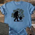 Squatch BBQ Smoker Comfort Colors T-shirt in Blue Jean, laid flat. A black Bigfoot silhouette walks past a kettle grill on wheels, set against dark green palm tree silhouettes; no words are printed. The shirt lies on weathered wood beside olive pants and brown hiking boots.