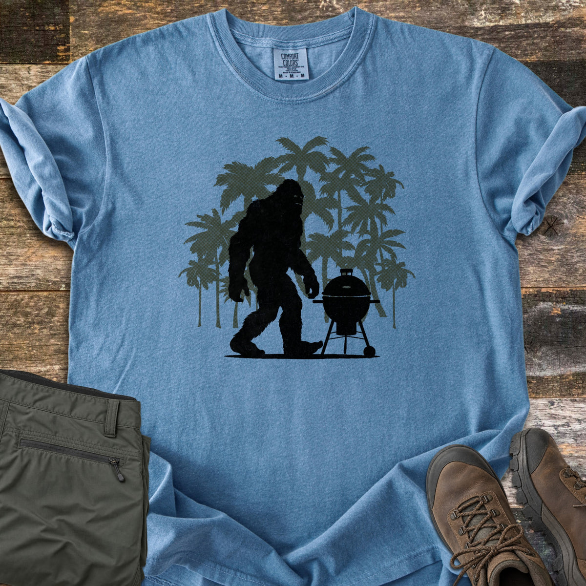 Squatch BBQ Smoker Comfort Colors T-shirt in Blue Jean, laid flat. A black Bigfoot silhouette walks past a kettle grill on wheels, set against dark green palm tree silhouettes; no words are printed. The shirt lies on weathered wood beside olive pants and brown hiking boots.