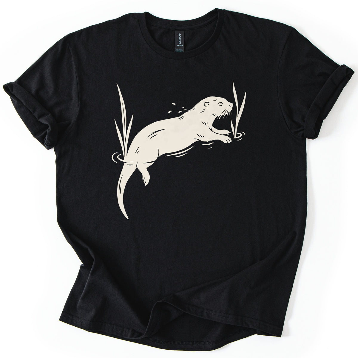 Swimming Otter Comfort Colors T-shirt in Black, laid flat. A cream linocut-style otter swims through rippling water between tall reeds, with droplets above its head. No words are printed. The sleeves are cuffed and the shirt lies on a white background.