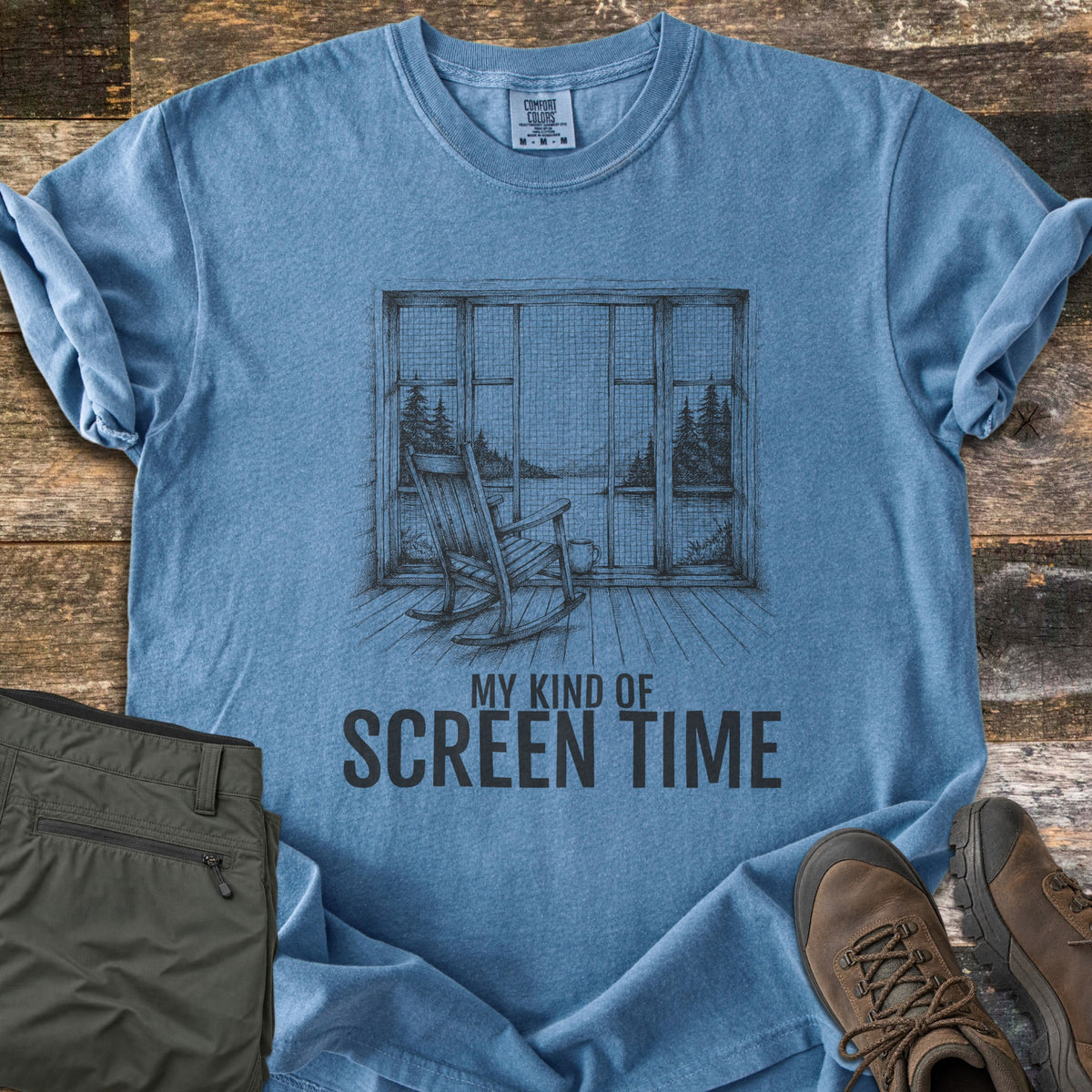 My Kind Of Screen Time Comfort Colors T-shirt in Blue Jean, laid flat. A pencil-style drawing of a wooden rocking chair and mug before a screened window showing a lake and pine trees, above the words My Kind Of Screen Time. Olive hiking pants and brown hiking boots rest on old wood planks.