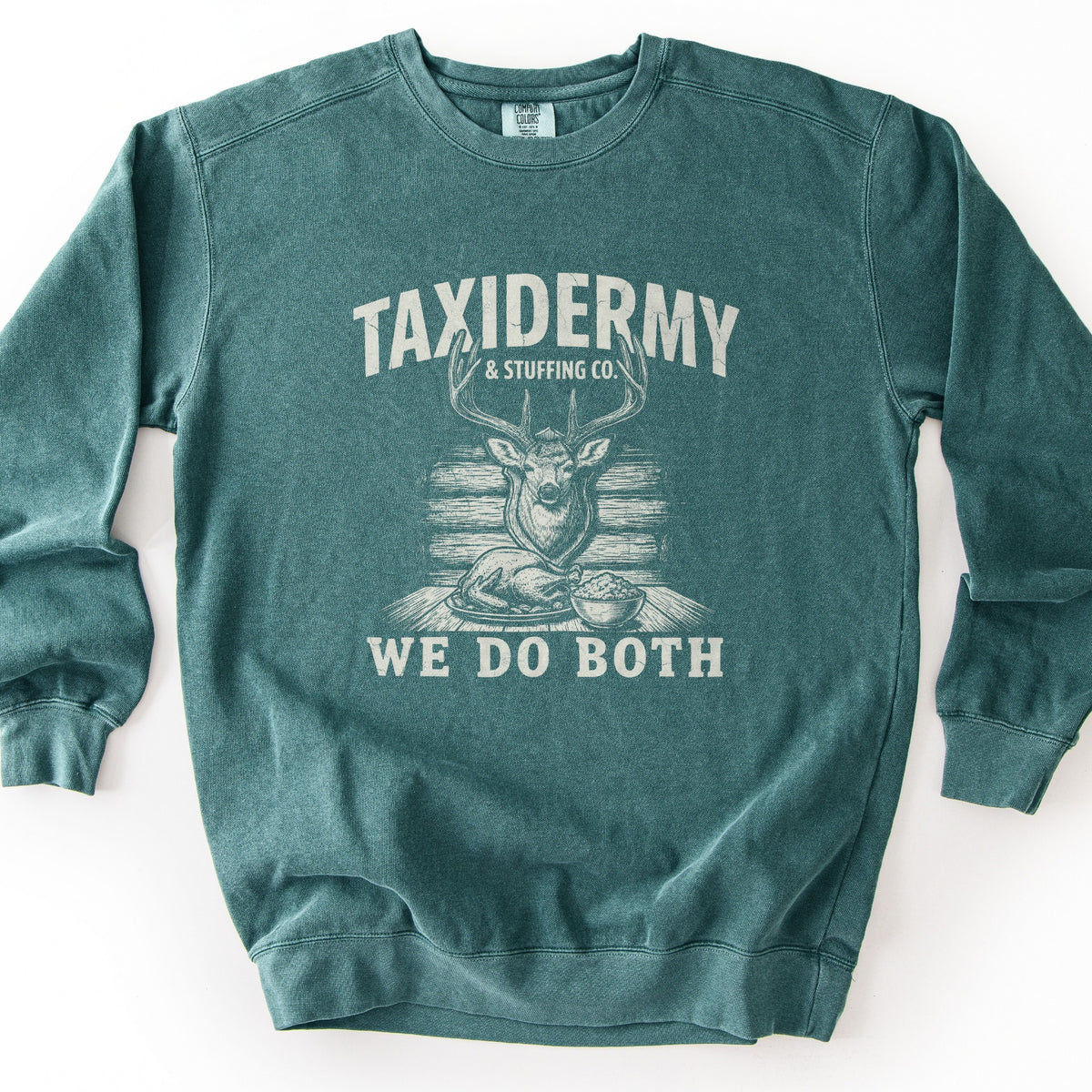 Taxidermy & Stuffing Co. We Do Both Comfort Colors crewneck sweatshirt in Blue Spruce, laid flat. A distressed cream print shows a mounted deer head above a roast turkey and a bowl of stuffing on a table, with the words Taxidermy & Stuffing Co. above and We Do Both below. Shown on a white background.