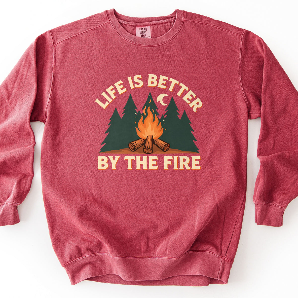 Life Is Better By The Fire Comfort Colors crewneck sweatshirt in Crimson, laid flat. Cream letters with an orange shadow arch Life Is Better over dark green pine trees, a crescent moon and a campfire of crossed logs, with By The Fire below. It lies on a white surface.