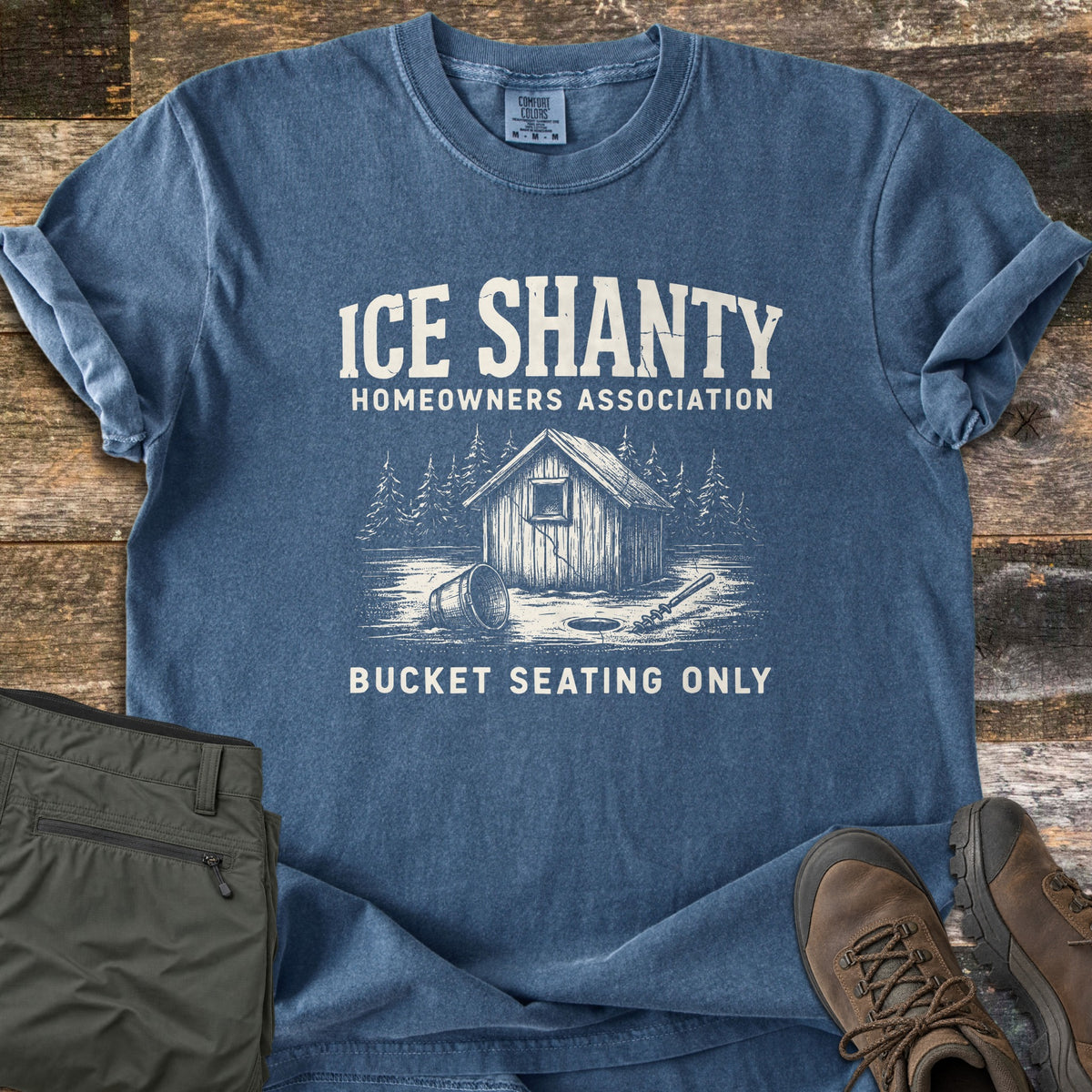 Ice Shanty Homeowners Association Comfort Colors T-shirt in Denim, laid flat. Cream print of a wooden ice shanty among pine trees, a tipped bucket, ice hole and auger, under Ice Shanty Homeowners Association and above Bucket Seating Only. Shown on wood with olive pants and brown boots.