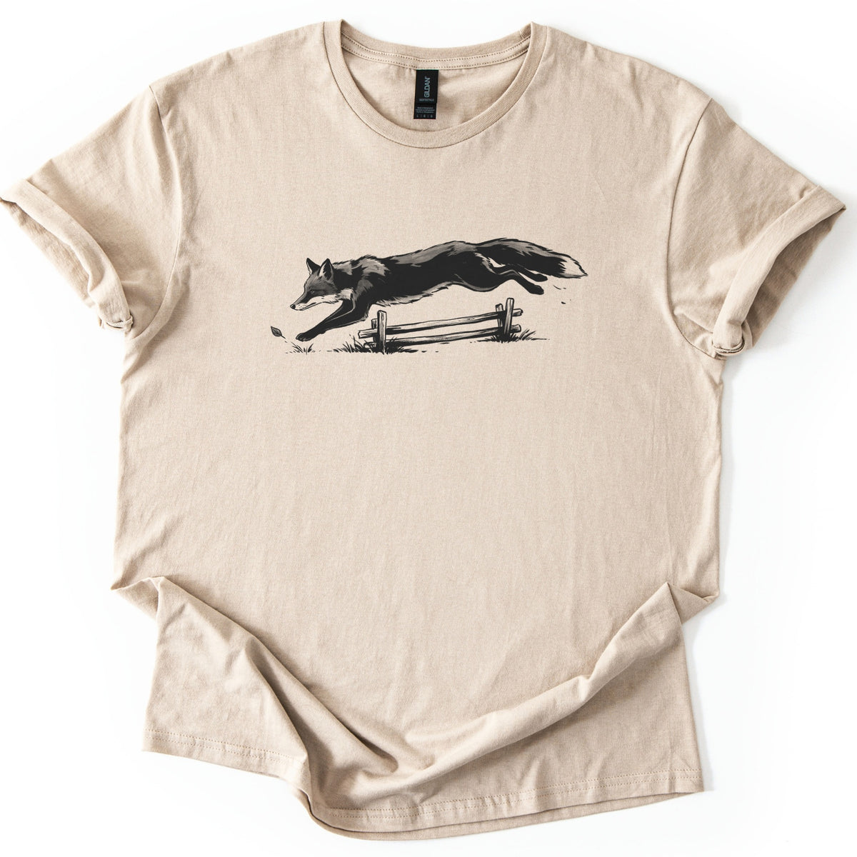 Leaping Fox Comfort Colors T-shirt in Sand, laid flat. A black and gray sketch of a fox leaping over a low wooden rail fence, with grass tufts and a falling leaf below. The shirt has cuffed sleeves and lies on a white background.