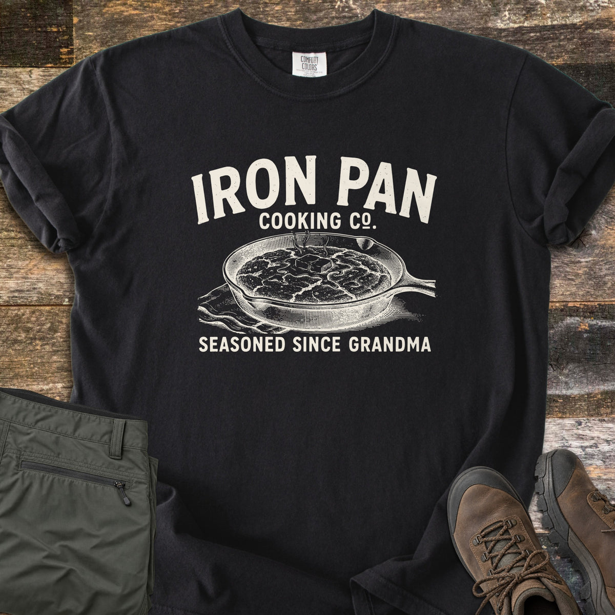 Iron Pan Cooking Co Comfort Colors T-shirt in Black, laid flat. Cream print of a cast iron skillet of cornbread with melting butter on a napkin, under the words Iron Pan Cooking Co. and above Seasoned Since Grandma. Folded olive pants and brown hiking boots lie on rustic wood.