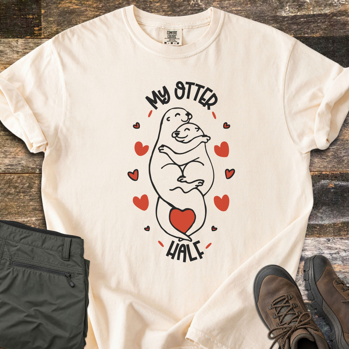 My Otter Half Comfort Colors T-shirt in Ivory, laid flat. Two smiling cartoon otters hug in black line art, ringed by red hearts with a red heart at their tails, between the words My Otter Half. Folded olive pants and brown hiking boots sit beside it on weathered wood.
