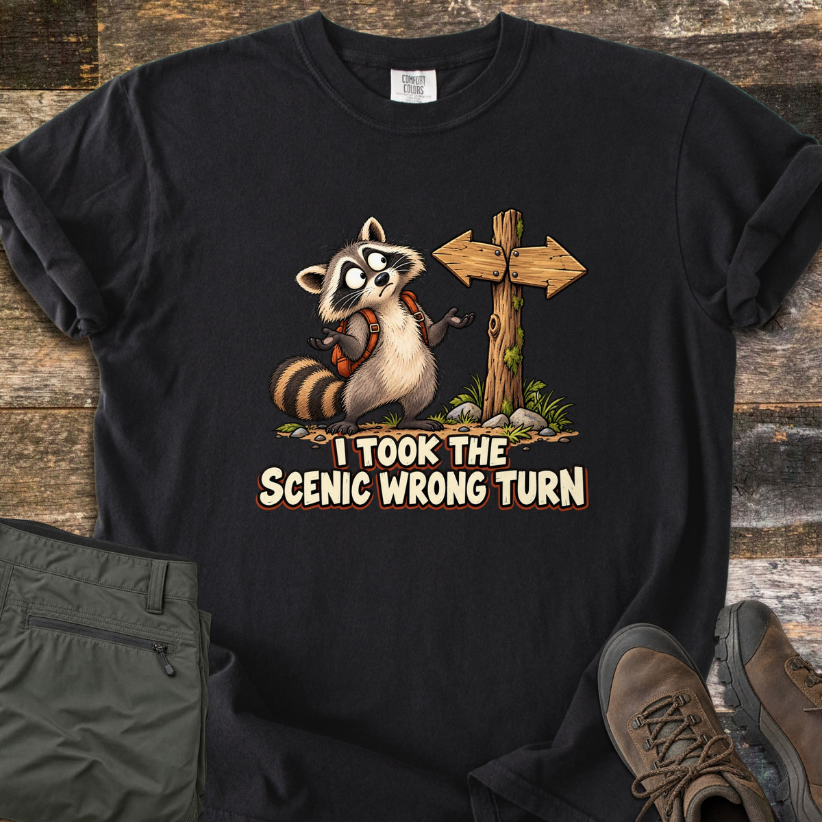 I Took the Scenic Wrong Turn Comfort Colors T-shirt in Black, laid flat. A cartoon raccoon with a backpack shrugs, looking confused beside a wooden signpost with arrows pointing both ways, above the words I Took the Scenic Wrong Turn. Olive hiking pants and brown boots lie on weathered wood.