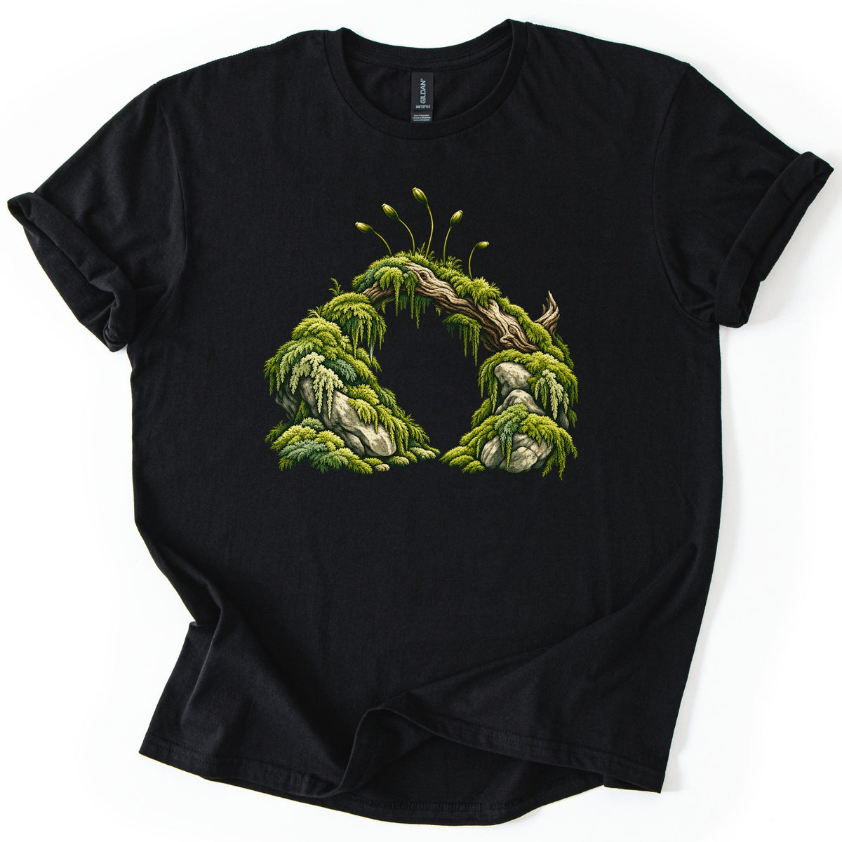 Moss Arch Comfort Colors T-shirt in Black, laid flat. A green moss-draped log arches between two mossy boulders, with thin spore stalks rising from the top. No words are printed. The shirt lies on a white background with its sleeves rolled.