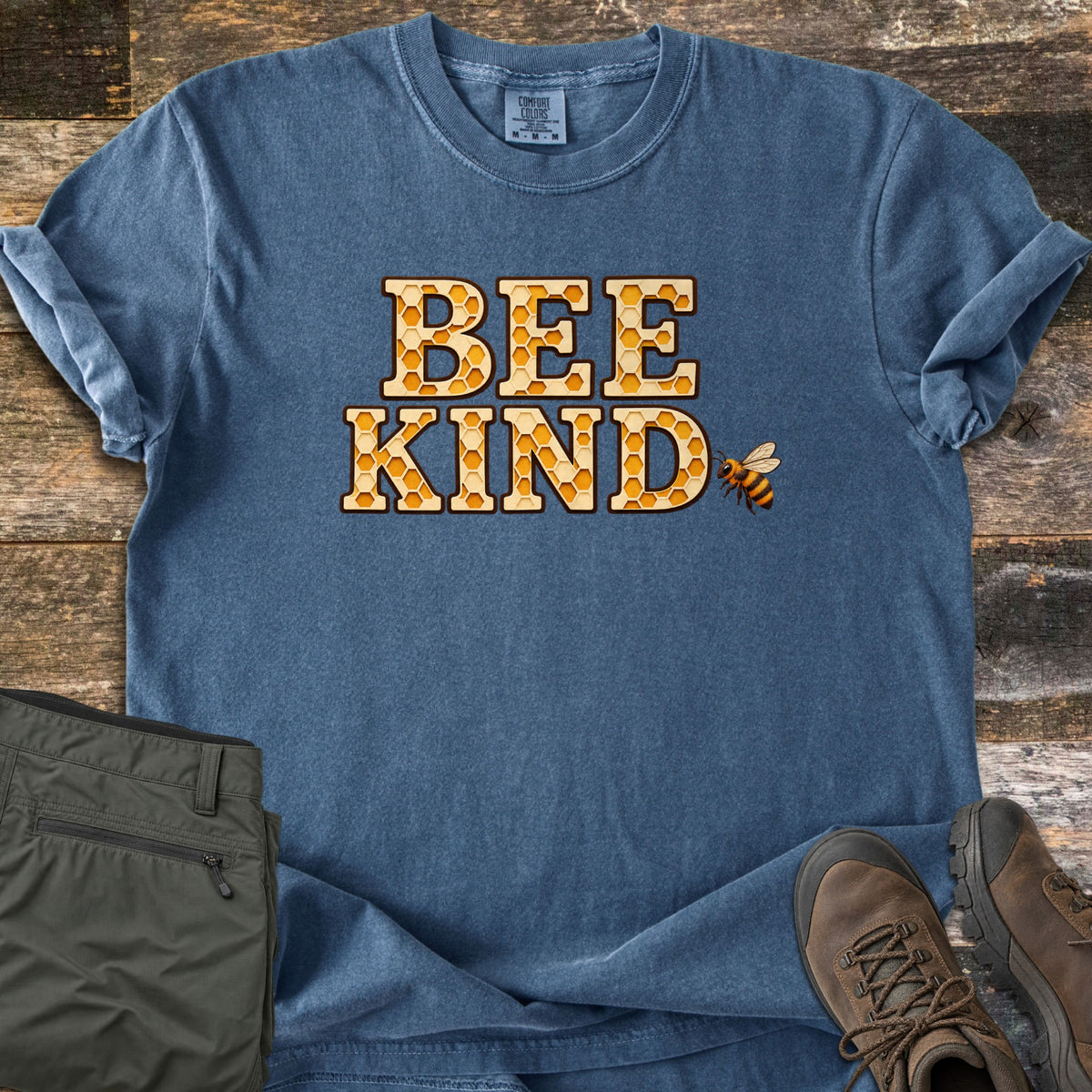 Bee Kind Comfort Colors T-shirt in Denim, laid flat. The words BEE KIND in tall letters filled with a golden honeycomb pattern, with a small honeybee beside the D. Sleeves are rolled on a wood plank surface next to olive pants and brown hiking boots.