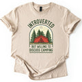 Introverted Camping Comfort Colors T-shirt in Sand, laid flat. An arched badge shows a red tent among green pine trees under a pale moon, with INTROVERTED curved above and BUT WILLING TO DISCUSS CAMPING below in dark green. The shirt lies on a white background.