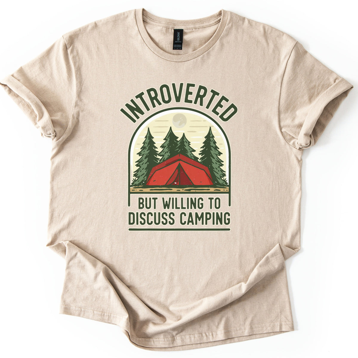 Introverted Camping Comfort Colors T-shirt in Sand, laid flat. An arched badge shows a red tent among green pine trees under a pale moon, with INTROVERTED curved above and BUT WILLING TO DISCUSS CAMPING below in dark green. The shirt lies on a white background.