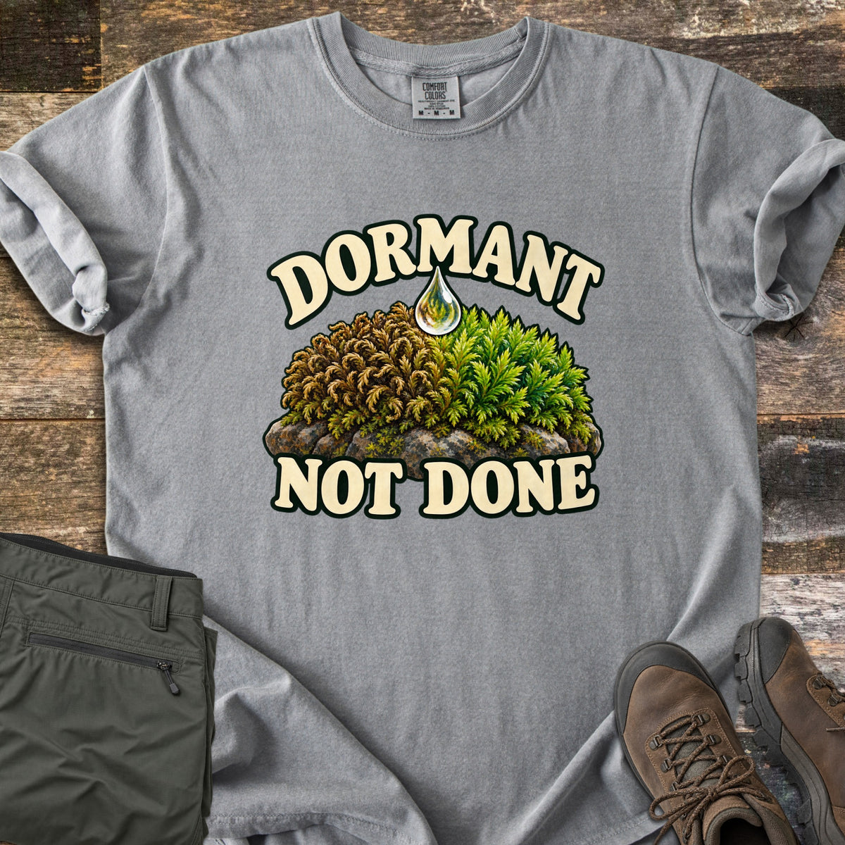 Dormant Not Done Comfort Colors T-shirt in Grey, laid flat. Dormant is printed in cream over a moss mound on a rock, brown and curled on the left and green on the right, under a water drop, with Not Done below. Olive pants and brown hiking boots sit on weathered wood.