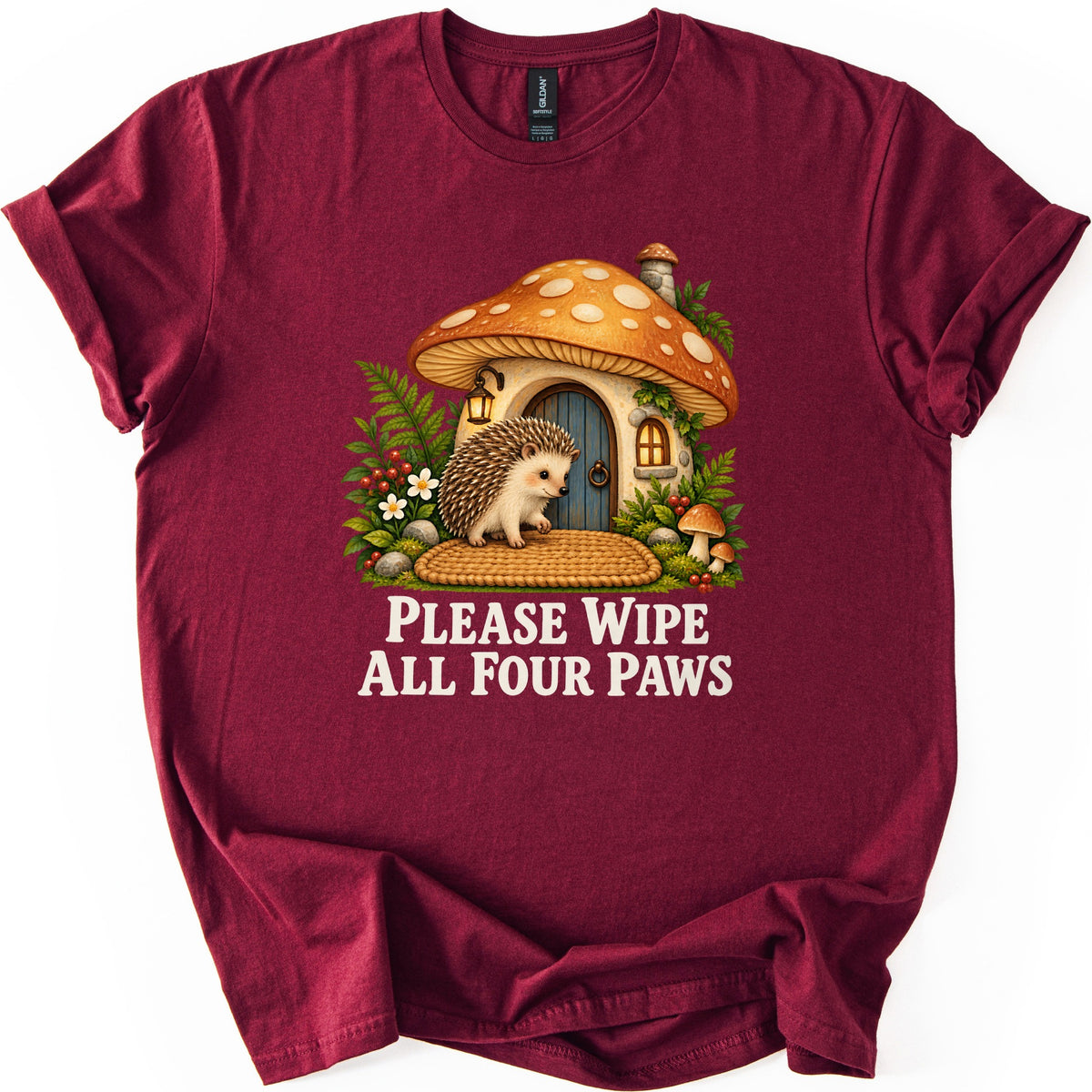 Please Wipe All Four Paws Comfort Colors T-shirt in Maroon, laid flat. A hedgehog on a woven doormat waits at the blue door of a mushroom cottage with an orange spotted cap, ferns, white flowers and red berries. Below, cream text reads Please Wipe All Four Paws.