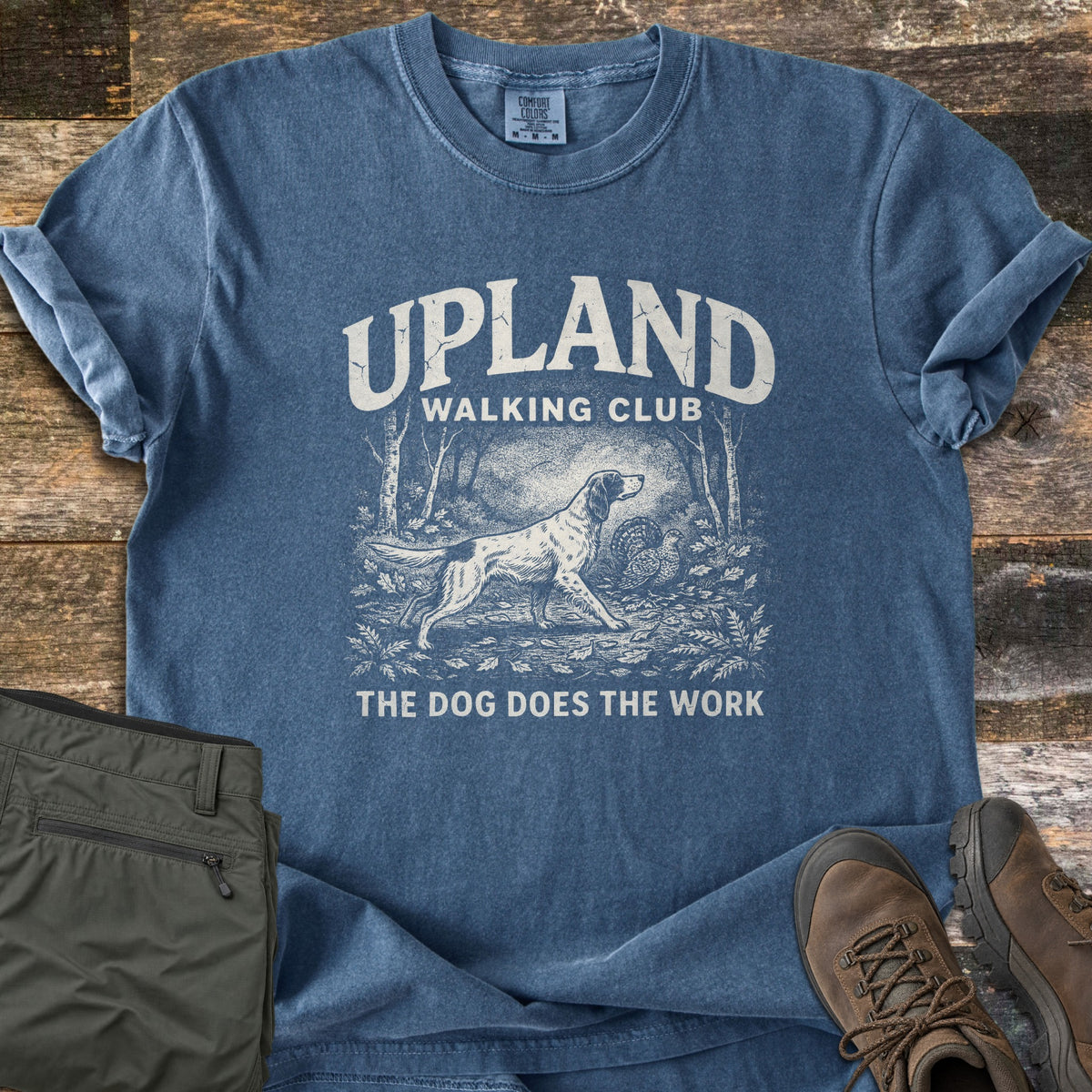 Upland Walking Club Comfort Colors T-shirt in Denim, laid flat. A spotted setter dog stalks a grouse in a wooded scene, under UPLAND WALKING CLUB and above THE DOG DOES THE WORK. Olive hiking pants and brown boots sit beside it on weathered wood.
