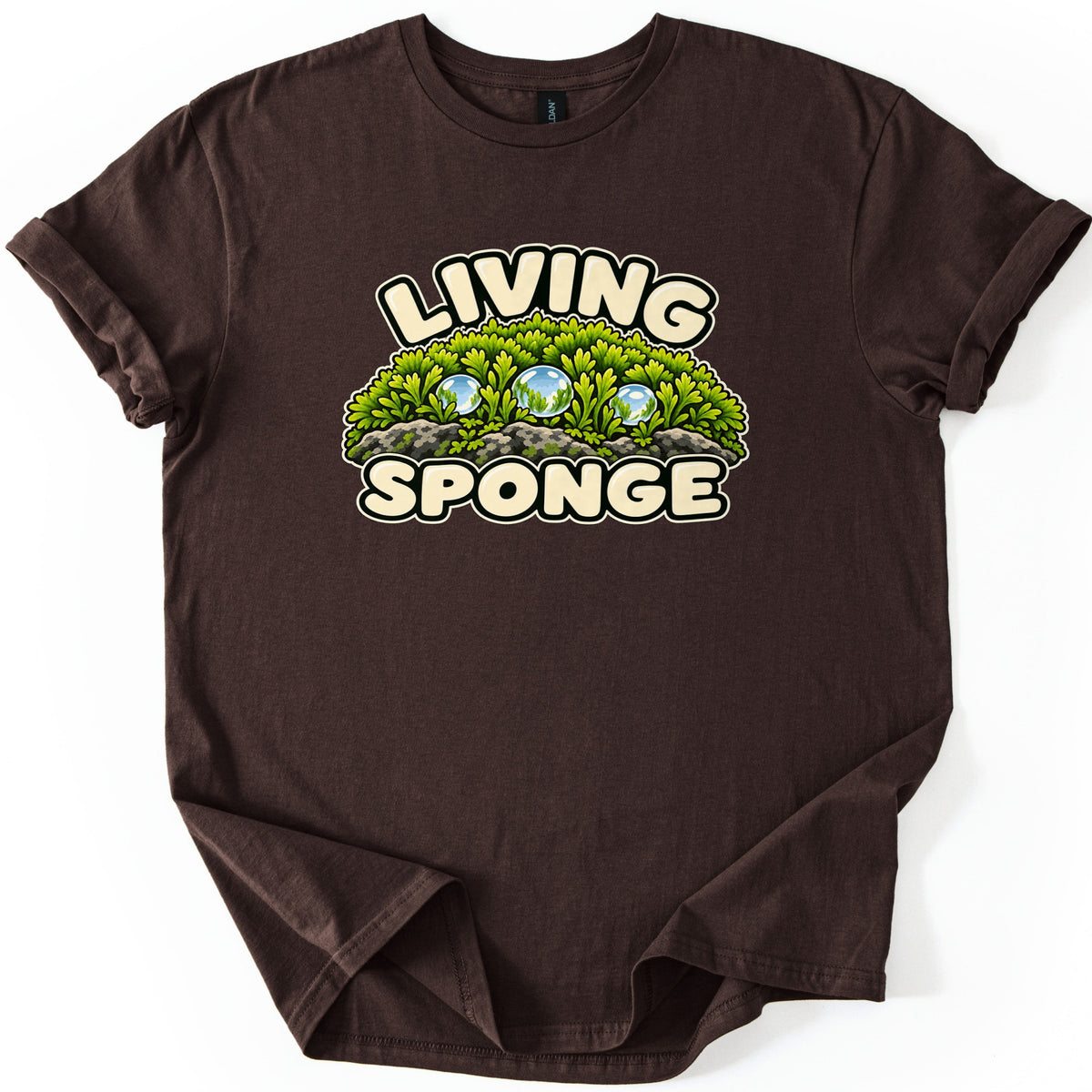 Living Sponge Comfort Colors T-shirt in Dark Chocolate, laid flat. The words LIVING and SPONGE in cream bubble letters frame a cartoon mound of green moss on gray rocks dotted with three clear water droplets. The shirt has rolled sleeves on a white background.