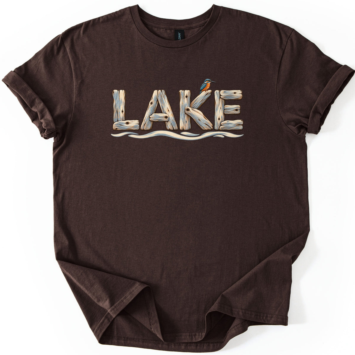 Lake Comfort Colors T-shirt in Dark Chocolate, laid flat. The word LAKE is spelled in weathered driftwood planks, with a small blue and orange kingfisher perched on the K and wavy water lines below. The shirt lies on white with rolled sleeves and a folded hem.