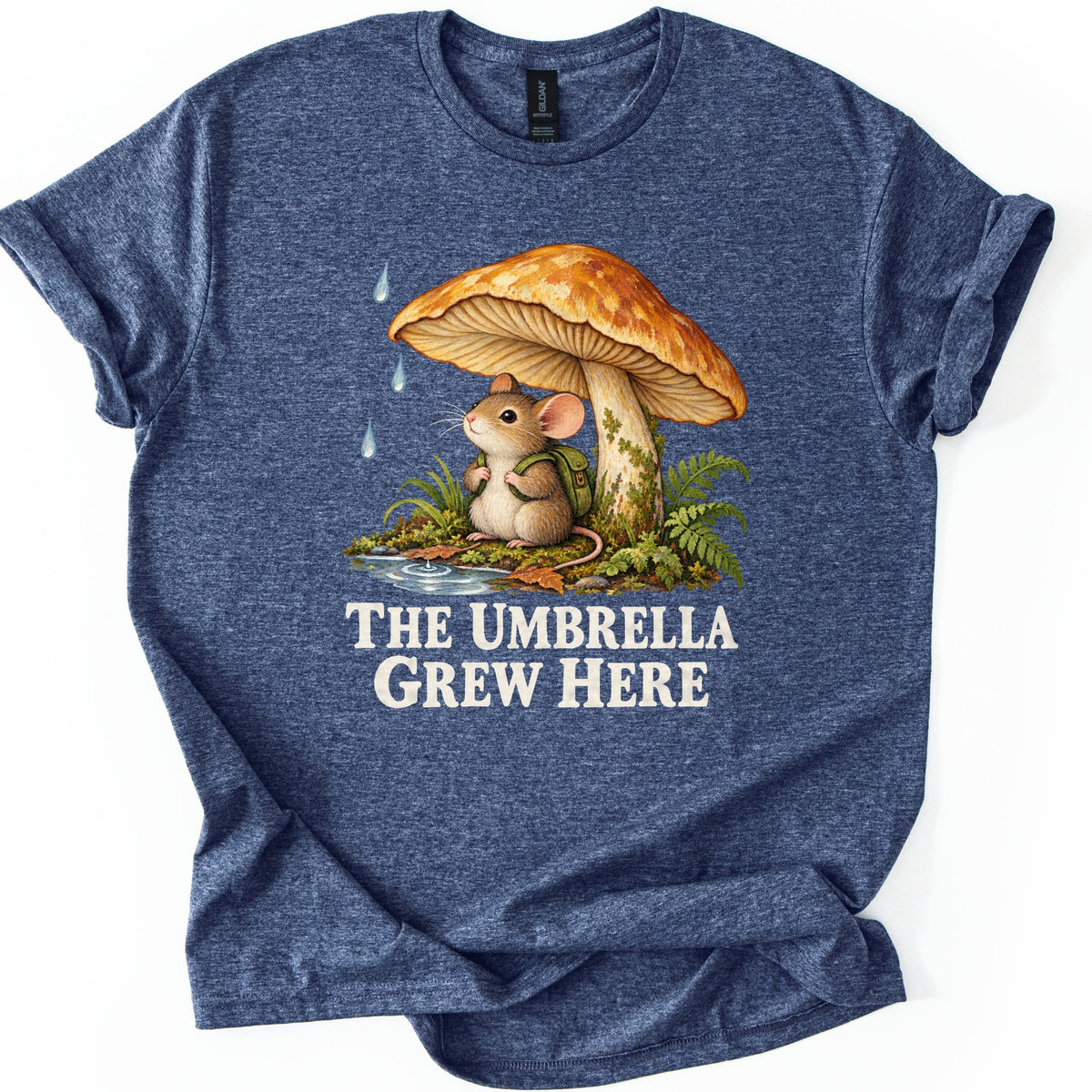 The Umbrella Grew Here Comfort Colors T-shirt in Heather Navy, laid flat. A mouse wearing a green backpack sits under a large orange mushroom as raindrops fall, beside ferns and a puddle, above the words The Umbrella Grew Here in cream letters. Shown on a white background.