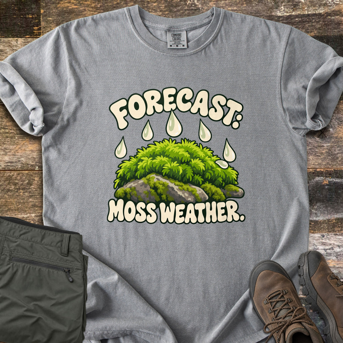 Forecast Moss Weather Comfort Colors T-shirt in Grey, laid flat. A cartoon mound of bright green moss on a gray rock, with raindrops falling from the words Forecast: above and Moss Weather. below. Olive hiking pants and brown hiking boots sit beside it on weathered wood boards.