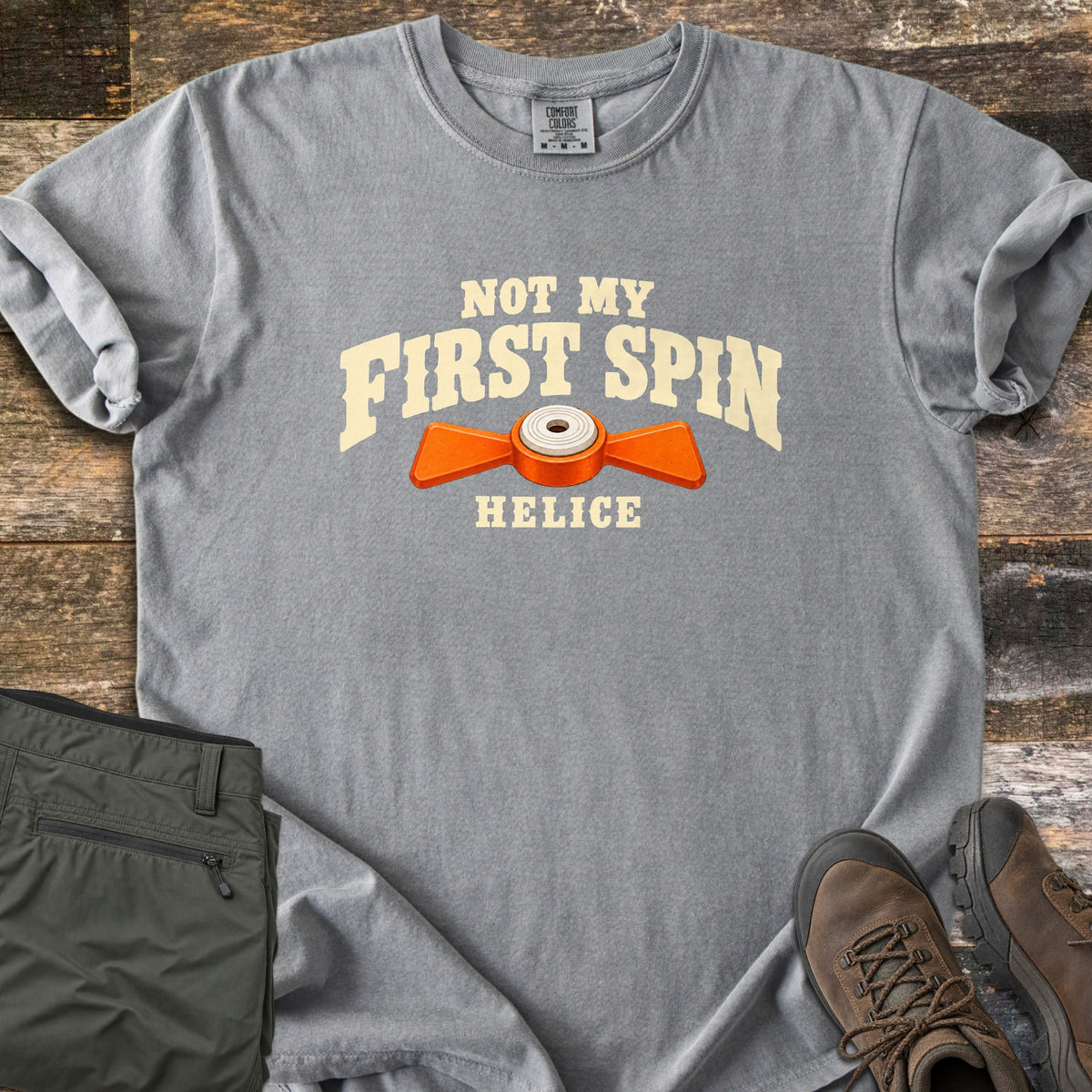 Not My First Spin Comfort Colors T-shirt in Grey, laid flat. Cream arched text reads Not My First Spin above an orange two-winged helice target with a white center, with Helice below. Folded olive pants and brown hiking boots sit beside it on weathered wood planks.