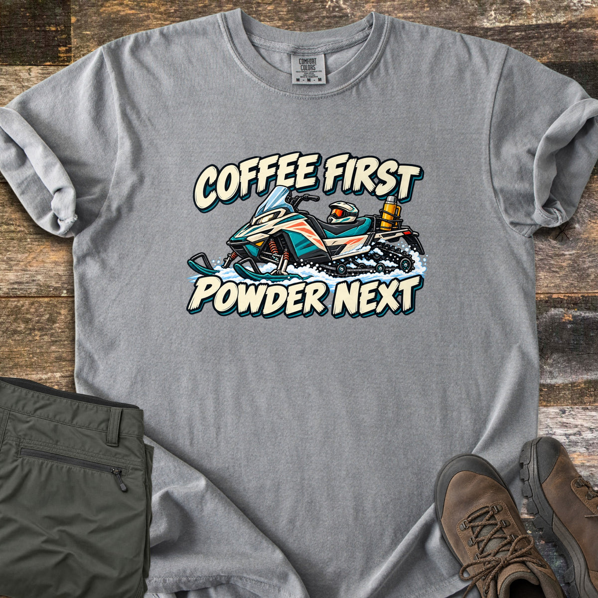 Coffee First Powder Next Comfort Colors T-shirt in Grey, laid flat. A cartoon snowmobile with a helmet on the seat and a yellow thermos strapped on back kicks up snow between the words Coffee First Powder Next. Olive hiking pants and brown boots sit on a wood floor.