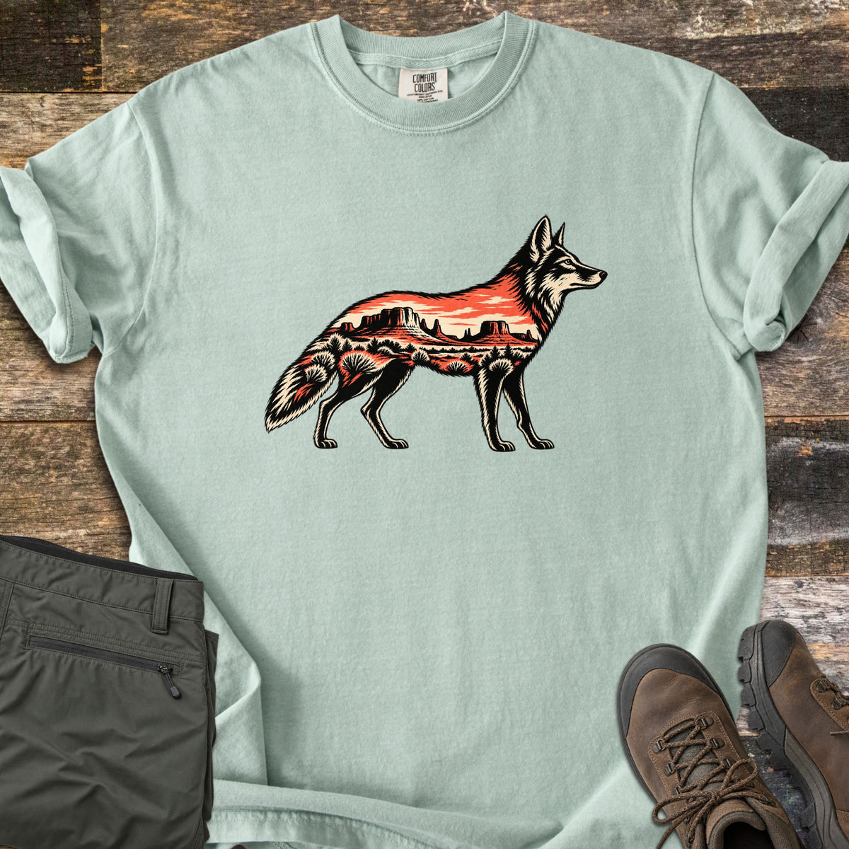 Desert Mesa Coyote Comfort Colors T-shirt in Bay, laid flat. A side-view coyote silhouette filled with a red desert mesa landscape, sunset sky and shrubs, with no words. Sleeves rolled on rustic wood, beside folded olive hiking pants and brown hiking boots.
