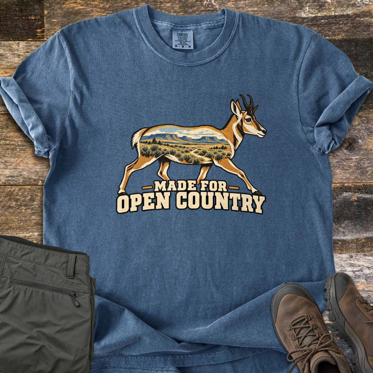 Made for Open Country Comfort Colors T-shirt in Denim, laid flat. A running pronghorn whose body is filled with a mesa and sagebrush landscape, above the words Made for Open Country. Sleeves cuffed, on weathered wood with olive pants and brown hiking boots.