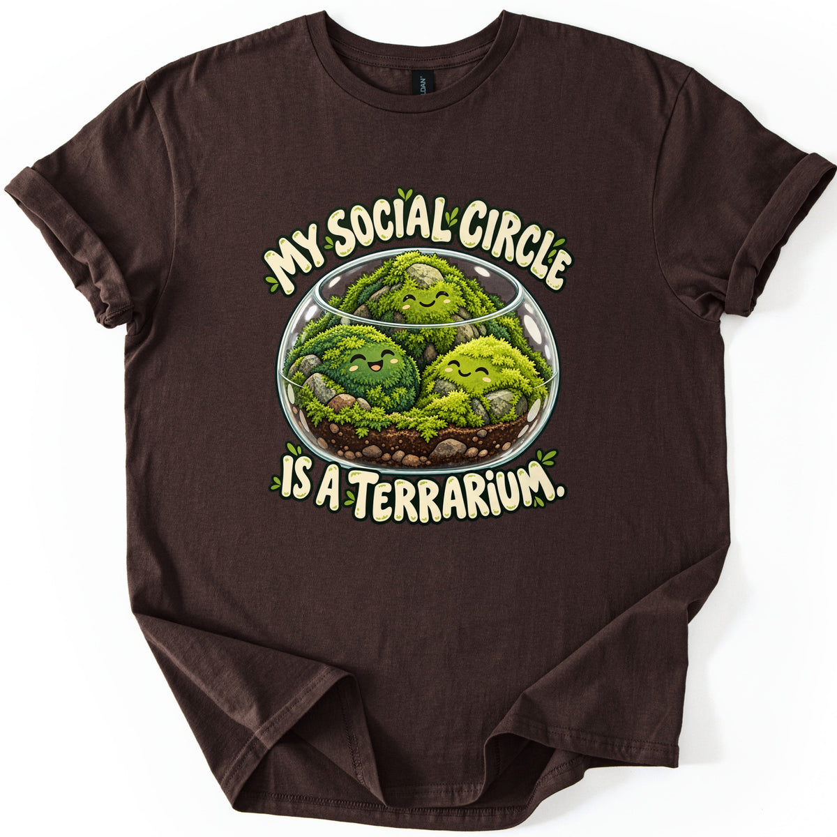 My Social Circle Is a Terrarium Comfort Colors T-shirt in Dark Chocolate, laid flat. A glass bowl terrarium with three smiling moss balls, stones and soil sits between the cream words My Social Circle and Is a Terrarium. The shirt lies flat on white with cuffed sleeves and a folded hem.