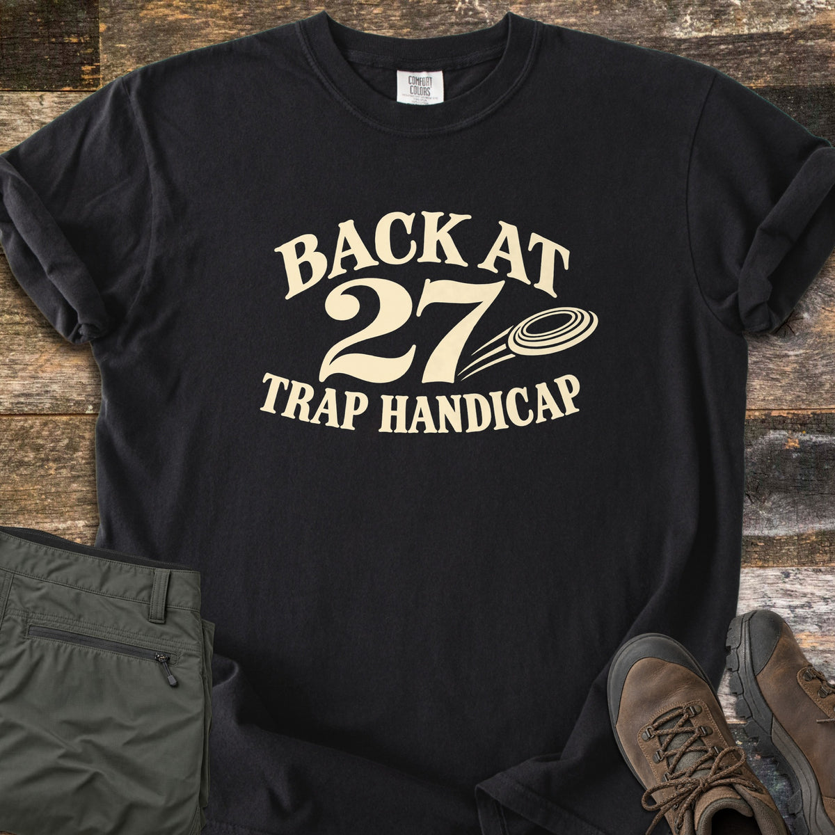 Back at 27 Trap Handicap Comfort Colors T-shirt in Black, laid flat. Cream print reads Back at 27 in an arch above Trap Handicap, with a clay target flying beside the 27. Sleeves rolled on a rustic wood surface, with olive cargo pants and brown hiking boots.